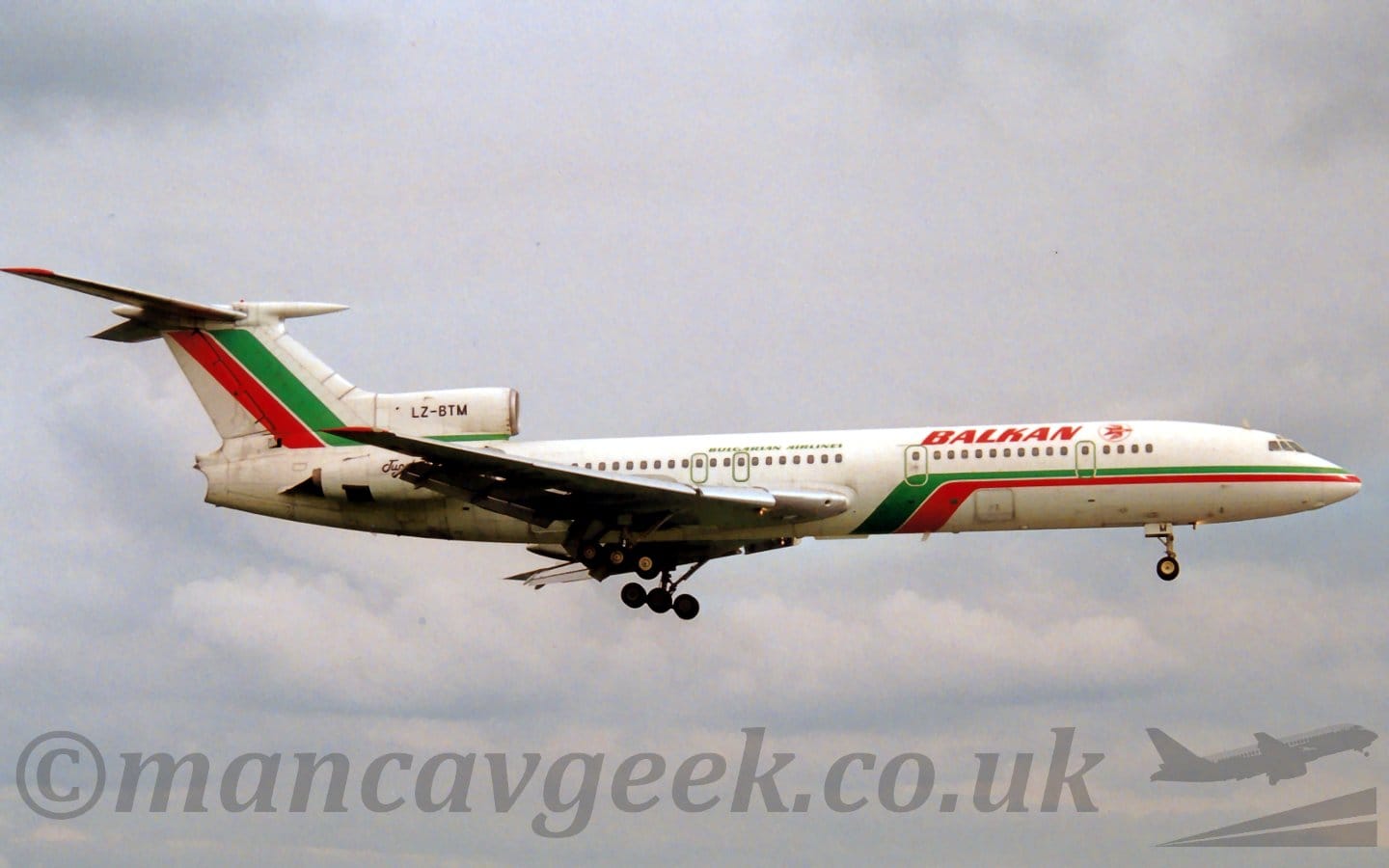 Side view of a 3 engined jet airliner flying from left to right at a low altitude, with undercarriage extended, and flaps deployed from the rear of the wings, suggesting it is about to land. The plane is mostly white, with a green and red stripe running along the body, sweeping under the belly just in front of the wings. There are red "Balkan" titles on the upper forward fuselage, next to the red outline of a circle containing a stylised red flying bird. There are smaller, green, "Bulgarian Airlines" on the upper fuselage, over the wings. There is a green and red diagonal stripe on the stripe, with the black registration "LZ-BTM" on the centre engine intake. The other engines are mounted on the sides of the rear fuselage, partially obscured by the wings. Cloudy grey sky fills the rest of the frame.