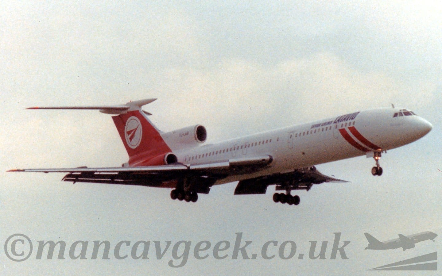 Side view of a 3 engined jet airliner flying from left to right at a low altitude, with undercarriage extended and flaps deployed from the rear of the wings, suggesting it is about to land. The plane is mostly white, with a pair of diagonal red bands around the lower forward fuselage, echoing the design of the Latvian flag. The rear fuselage, rear-mounted engines, and tail are red, with a white circle containing a stylise red flying bird on the tail. There is black text "Latvian Airlines" next to larger "LatAvio" titles on the upper forward fuselage, and the black registration on the white, centre engine intake at the front of the tail. Grey and white clouds with patches of blue threatening to break through fill the rest of the frame.