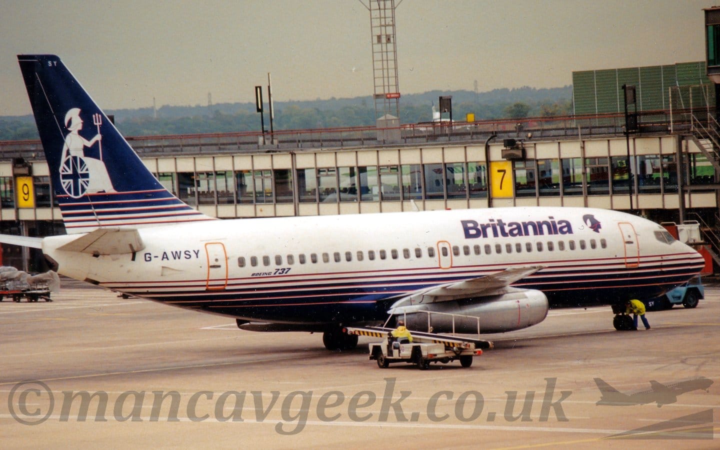 Side view of a twin engined jet airliner parked facing to the right. The plane is mostly white, with thin blue stripes on the lower fuselage getting wider until they merge into a solid block on the belly. There are dark blue "Britannia" titles on the upper forward fuselage, next to a head wearing an extravagantly plumed helmet. The tail is the reverse of the fuselage, white at the bottom, with thin blue lines getting thicker as they rise up the tail, merging into a solid mass at the top. A silhouetted, white, seated female figure wearing a robe and an elaborately plumed helmet, and holding a trident in her left hand and a white shield covered in a circular British flag in her right, down low by the ground, covers most of the rest of the tail, to modern eyes resembling a woman in a wheelchair. Grey concrete apron fills the foreground, with a baggage-loading truck driving up towards the rear of the plane. A terminal building stretches across the frame in the background, with tree covered hills in the distance, under leaden grey sky.