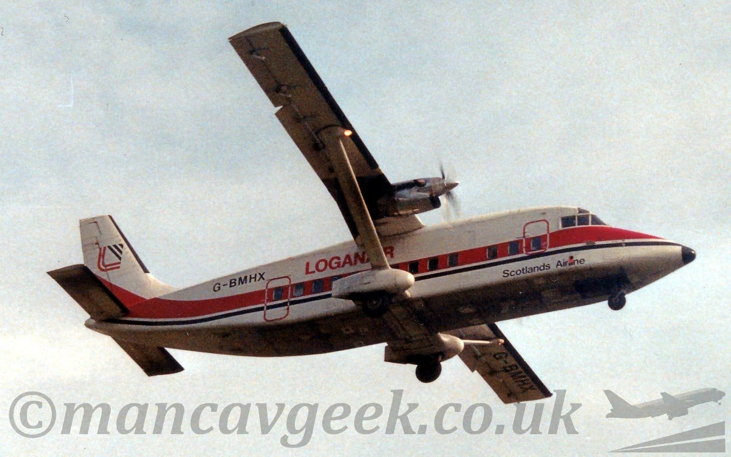 Low side view of a squat and boxy, high-winged, twin propellor-engined airliner flying from left to right at a low altitude, with undercarriage lowered and flaps extended from the rear of the wings, suggesting it is about to land. The plane is mostly white, with a thick red and thin black stripe running along the body, turning up in to the lower parts of the tail. The undercarriage is contained in sponsons attached by stub wings to the bottom of the fuselage, and by large diagonal supports to the mid-point of the wings. There are red "LoganAir" titles on the upper rear fuselage, the last few letters under the wings, , with the black registration "G-BMHX" further back on the upper rear fuselage, and black text "Scotlands Airline" on the lower forward fuselage, under the cockpit. The registration is also repeated under the outer panels of the left wing. The tail has a red and black logo, a pair of red lines bent through a right angle forming the left and bottom sides of a square, and 3 diagonal black lines of varying lengths stacked to form a triangle forming the other 2 sides. Flat grey sky fills the rest of the frame.