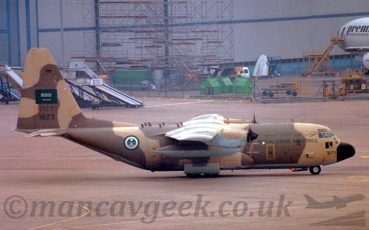 Side view of a high-winged, 4 propellor-engined military transport plane taxiing from left to right. The plane is covered in a dark brown and sandy brown camouflage scheme, with a grey belly. There are green "Royal Saudi Air Force" titles on the upper forward fuselage, in an Arabic script on top, and in English below, and the black number "1623" immediately behind the black nosecone. There is a green-white-green roundel on the rear fuselage, with white crossed swords over a white palm in the middle. The tail has a green fin flash in the middle, overlaid with some white text in a Arabic script, with the green Arabic number "1623" below that, and the English number below that. Grey concrete apron fills the foreground, and extends up to a large grey hangar in the distance, with the forward fuselage of a white jet airliner on the right, a large section of scaffolding in the middle, and a collection of mobile air-stairs on the left.