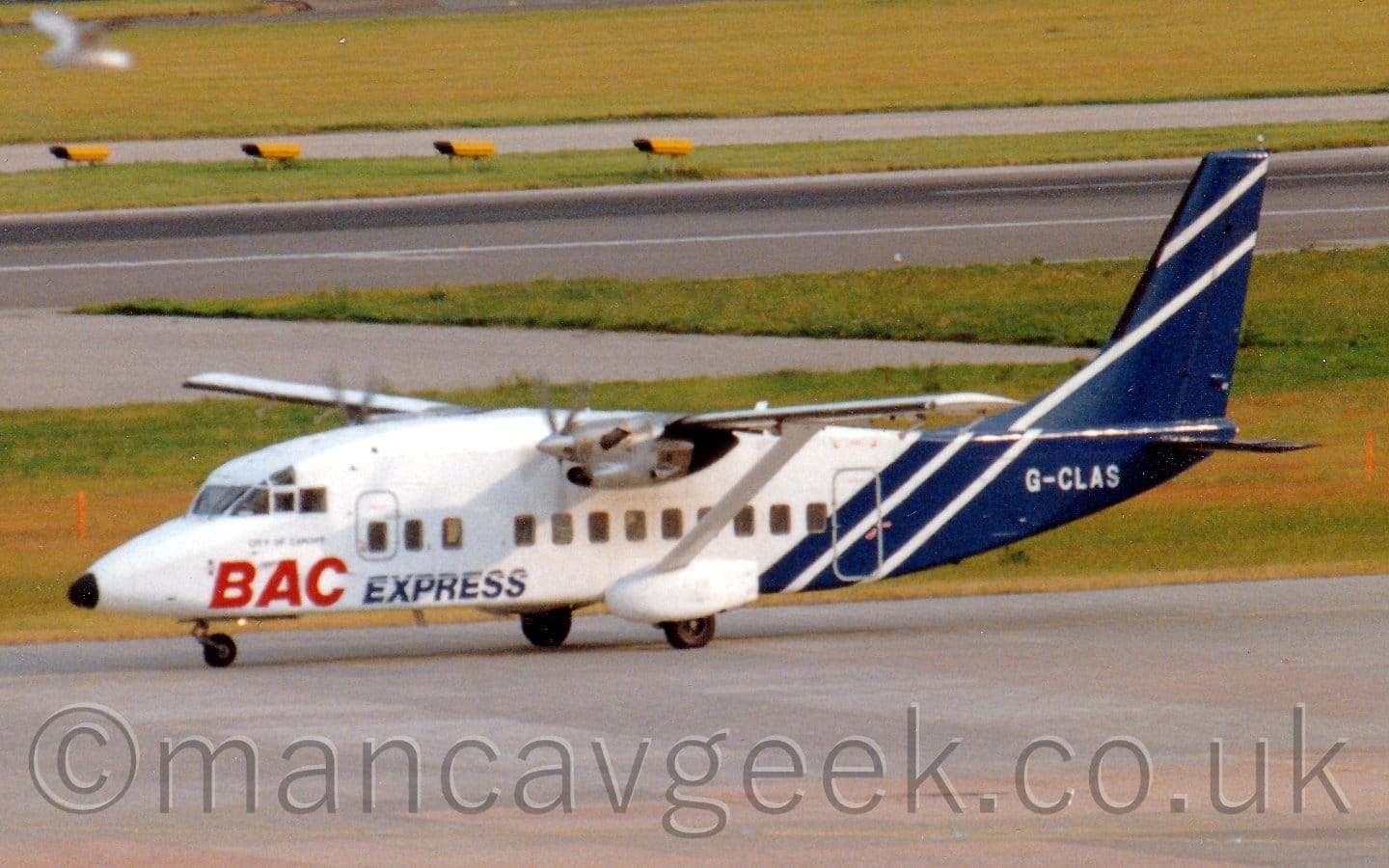 Side view of a boxy, high-winged, twin propellor-engined airliner taxiing from right to left. The plane is mostly white, with large red "BAC"  and smaller, dark blue "Express" titles on the lower forward fuselage, below the cockpit and forward cabin windows. The tail and rear fuselage are dark blue, with a pair of diagonal white stripes, and the white registration "G-CLAS". Grey concrete apron fills the foreground, with large grassed areas and a black runway filling the background.
