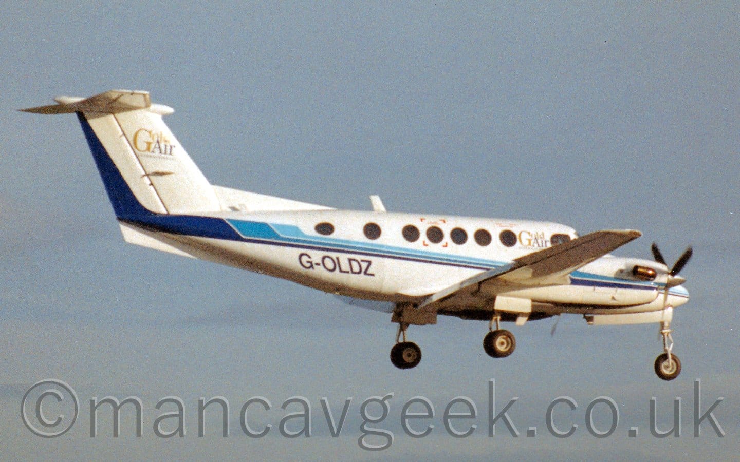 Side view of a twin propellor-engined BizProp flying from left to right at a low altitude, with undercarriage lowered, and flaps extended from the rear of the wings, suggesting it is about to land. The plane is mostly white, with a 3-tone blue stripe running along the body, gold and black "Gold Air" titles on the upper forward fuselage just aft of the cockpit windows, and the black registration "G-OLDZ" on the lower rear fuselage. A larger form of the gold and black "GoldAir" titles appears on the white tail. Bright sunlight reflects strongly off the plane, suggesting it is getting quite deep in to a summers evening. Hazy blue sky fills the background.