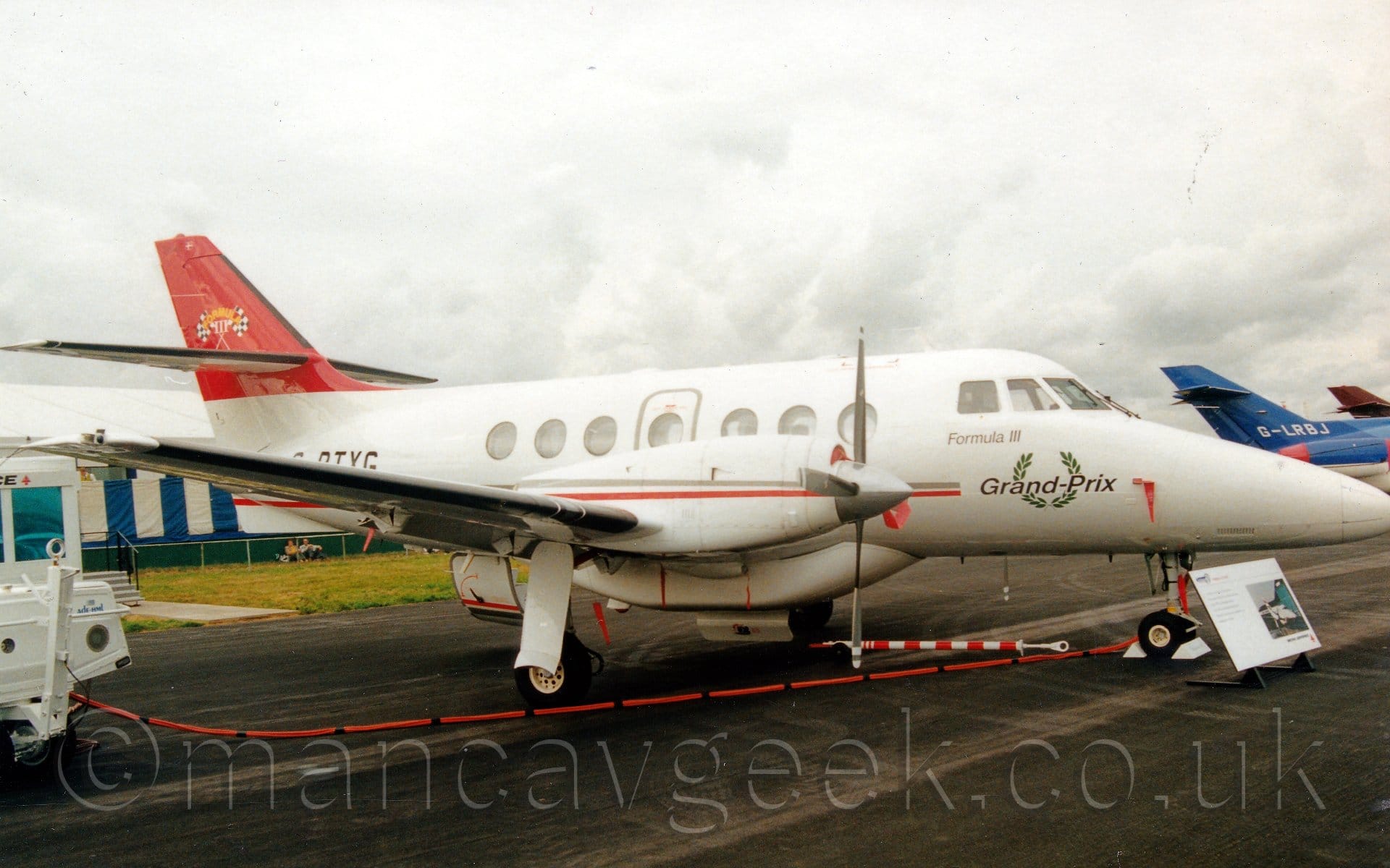 Side view of a twin propellor-engined airliner parked facing to the right on a black tarmac taxiway at an airshow. The plane is mostly white, with a grey and red stripe running along the body, as well as along the engine pods over the wings. There is a white baggage pod under the fuselage, covering the wing root. There is a green laurel wreathe on the nose, under the cockpit windows, , overlaid with the black words "Grand Prix", and the golden words "Formula III" directly under the cockpit windows. The tail is red, with a pair of crossed black and white checkered flags in the middle. There is a large board in front of the nosewheel, giving some information about the plane to onlookers. A thick red power cable runs from the nosewheel bay off to the left of the frame, behind a small white tow truck. The tails of a couple of planes - one blue, the other red - can be seen on the right of the frame, with cloudy grey sky filling the rest.
