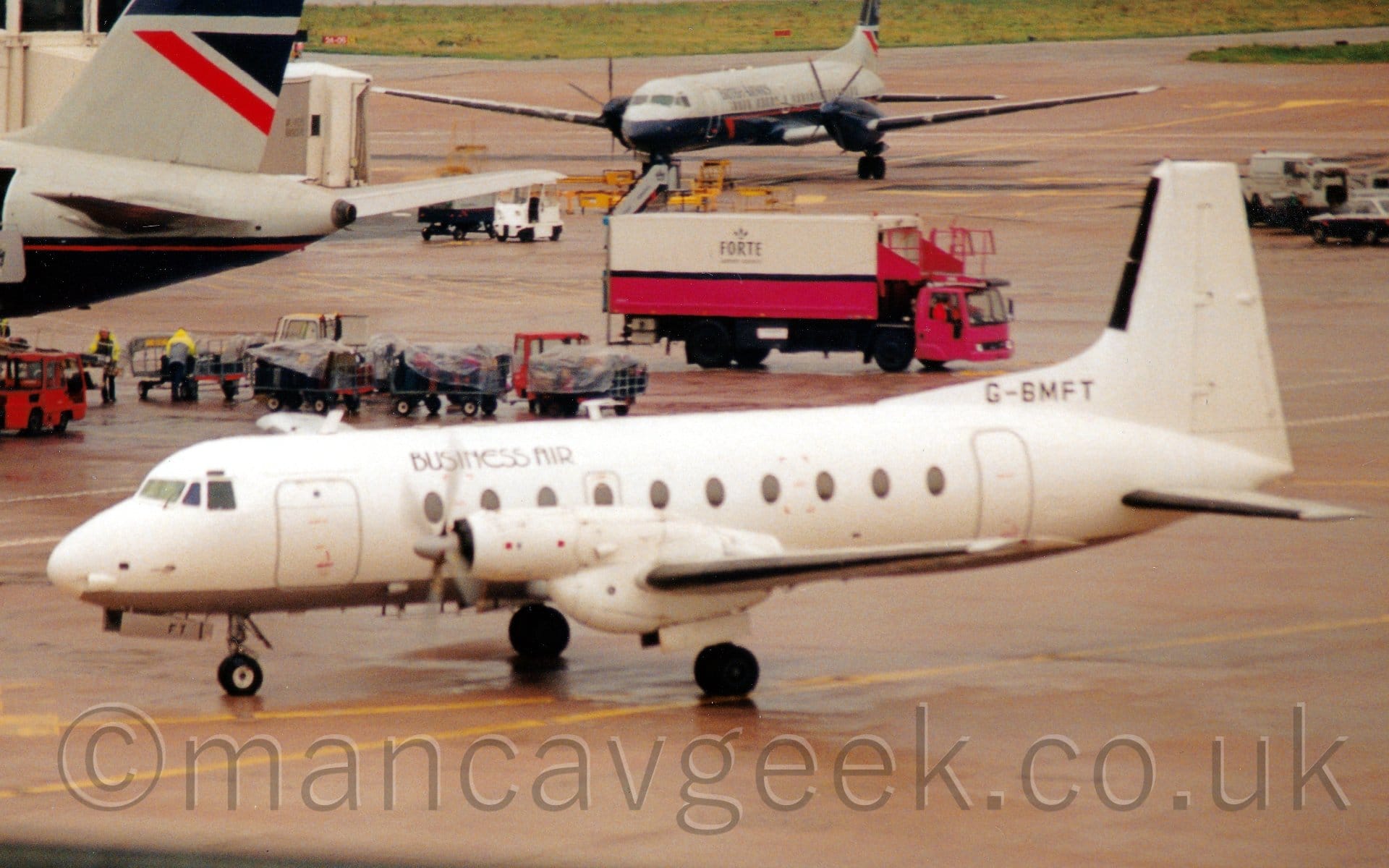 Side view of a twin propellor-engined airliner taxiing from right to left. The plane is almost entirely white, with a grey belly, and only small black "Business Air" titles on the upper forward fuselage, and the black registration "G-BMFT" on the upper rear marring the canvas. Grey concrete apron fills most of the frame, with the dark blue, grey, and red rear fuselage and tail of a much larger jet airliner poking into frame on the left of the background. A line of full baggage cart covered with plastic sheets stretches towards the middle of the frame from under the tail of that plane, with a white and pink catering truck with black "Forte" titles  beyond them, driving to the right. Another dark blue, grey, and red plane, this time with a propellor engine on each wing, sits right at the back, facing to the left.