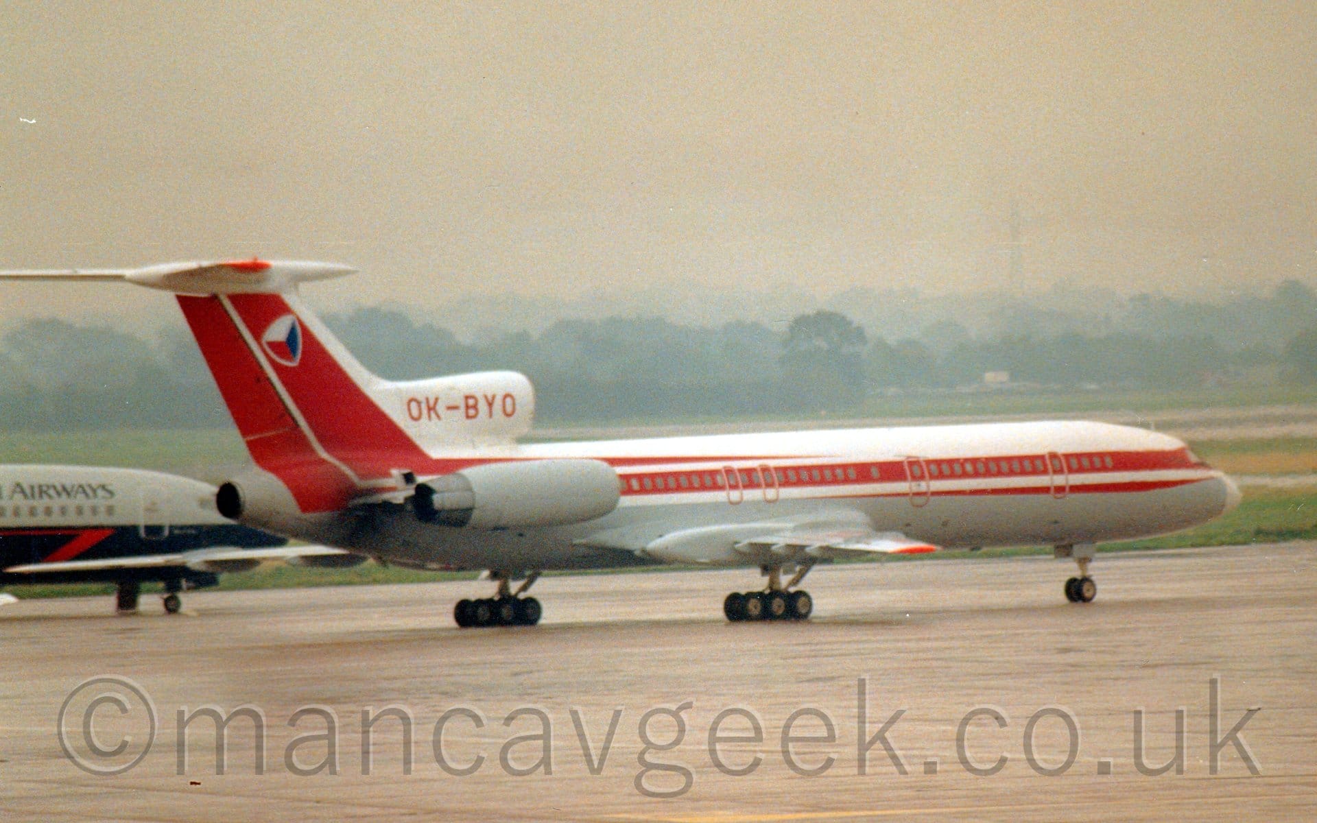 Side view of a 3 engined jet airliner, with the engines mounted on the rear fuselage, taxiing from left to right. The plane has white upper surfaces, with a grey belly, and a thick red stripe running along the body, covering the passenger cabin windows, before turning up to fill the tail. There is a slightly distorted red, blue, and white cube on the tail, and the red registration "OK-BYO" on the centre engine inlet above the rear fuselage. Grey concrete apron fills the foreground, with the forward fuselage of a grey and dark blue jet airliner in the background on the left. Trees in the distance stretch across the frame, slowly vanishing into hazy grey skies.
