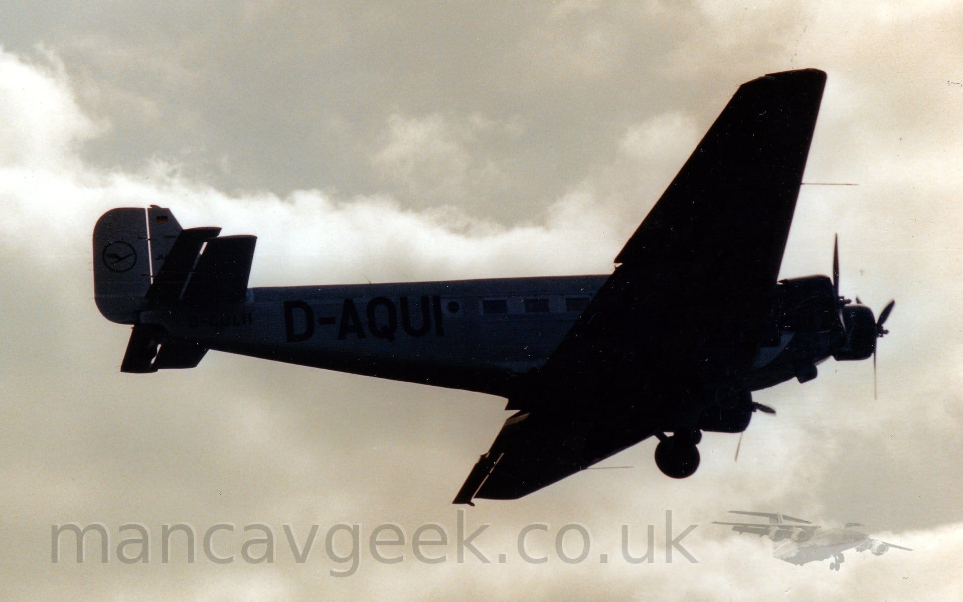Heavily backlit shot of a 3 propellor-engined airliner flying from left to right at a low altitude, with it's fixed tail-wheel undercarriage in place (obviously), and flaps extended from the rear of the wings, suggesting it is about to land. The fuselage is metallic grey, with a corrugated surface, and the black registration "D-AQUI" on the rear fuselage. Dramatic grey sky fill the background.