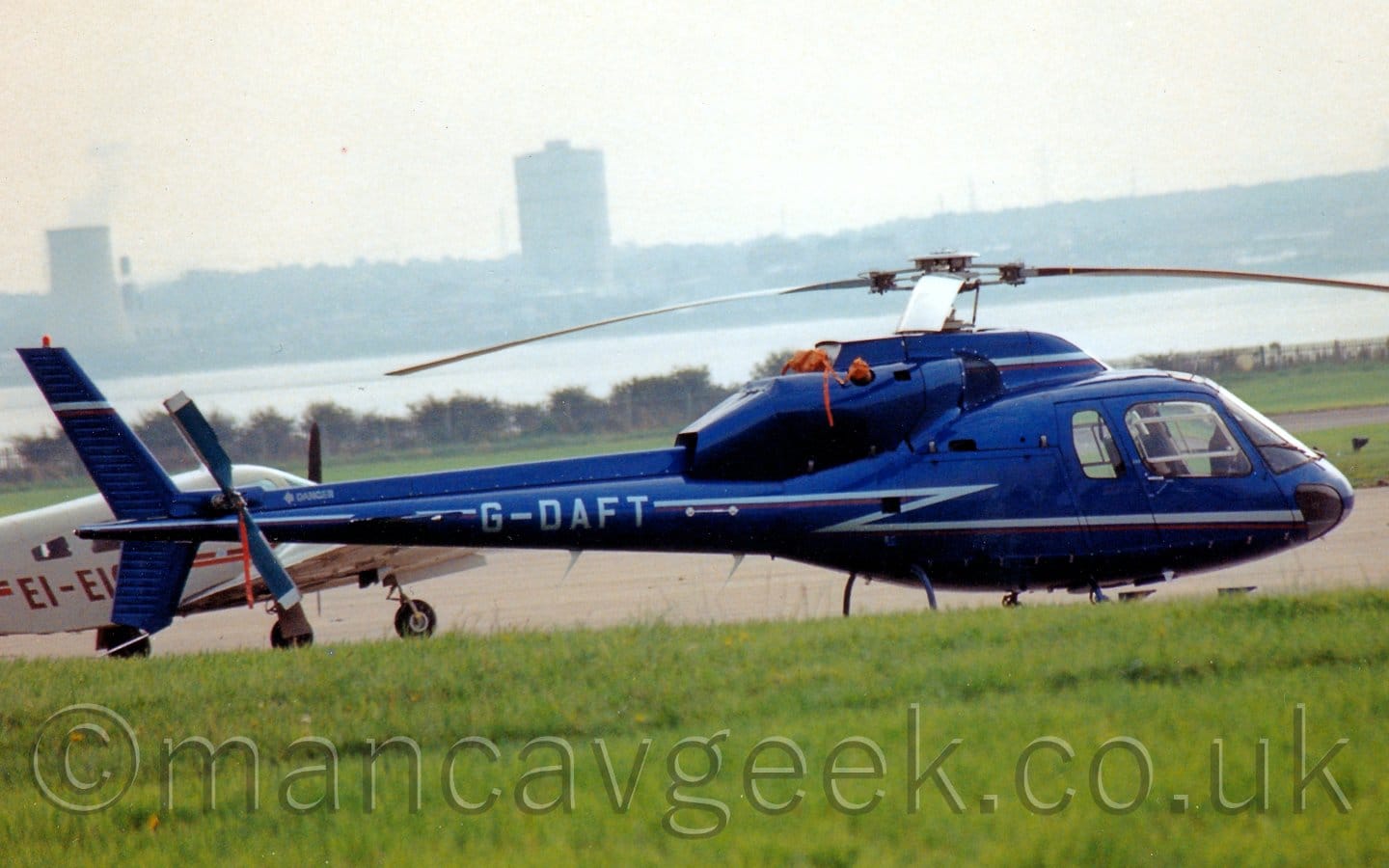 Side view of a helicopter parked a concrete apron area facing to the right. The chopper is cobalt blue, with a lighter blue stripe zig-zagging it's way across the body and tail-boom, overlaid with light blue registration "G-DAFT" on the boom. Another light blue stripe runs along the engine cowling mounted on top of the roof. Lush green grass fills the foreground, with a large grey apron area in the background. A brown, twin-engined light aircraft is parked behind this helicopter on the left. A wide river in the background has tall buildings on the far bank slowly vanishing into hazy grey sky.