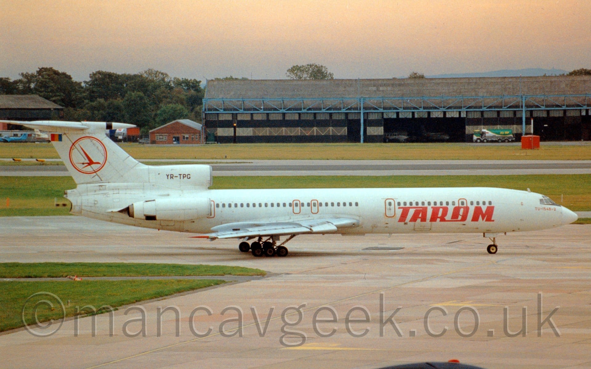 Side view of a 3 engined jet airliner, with the engines mounted on the rear fuselage, taxiing from left to right. The plane is almost entirely white, with red "Tarom" titles on the lower forward fuselage, the black registration "YR-TPG" on the centre engine inlet, and the outline of a red circle with a stylised red flying bird in the middle. Grey concrete apron fills the foreground, with a large grassed area on the left, with more grass lining the sides of a black runway in the background, with a wide black hangar with blue support beams stretching across the frame in the distance with a green and white fuel bowser parked in front, and several planes visible inside through the wide open doors. Trees in the distance on the left and slightly orange tinged sky fills the rest of the frame.