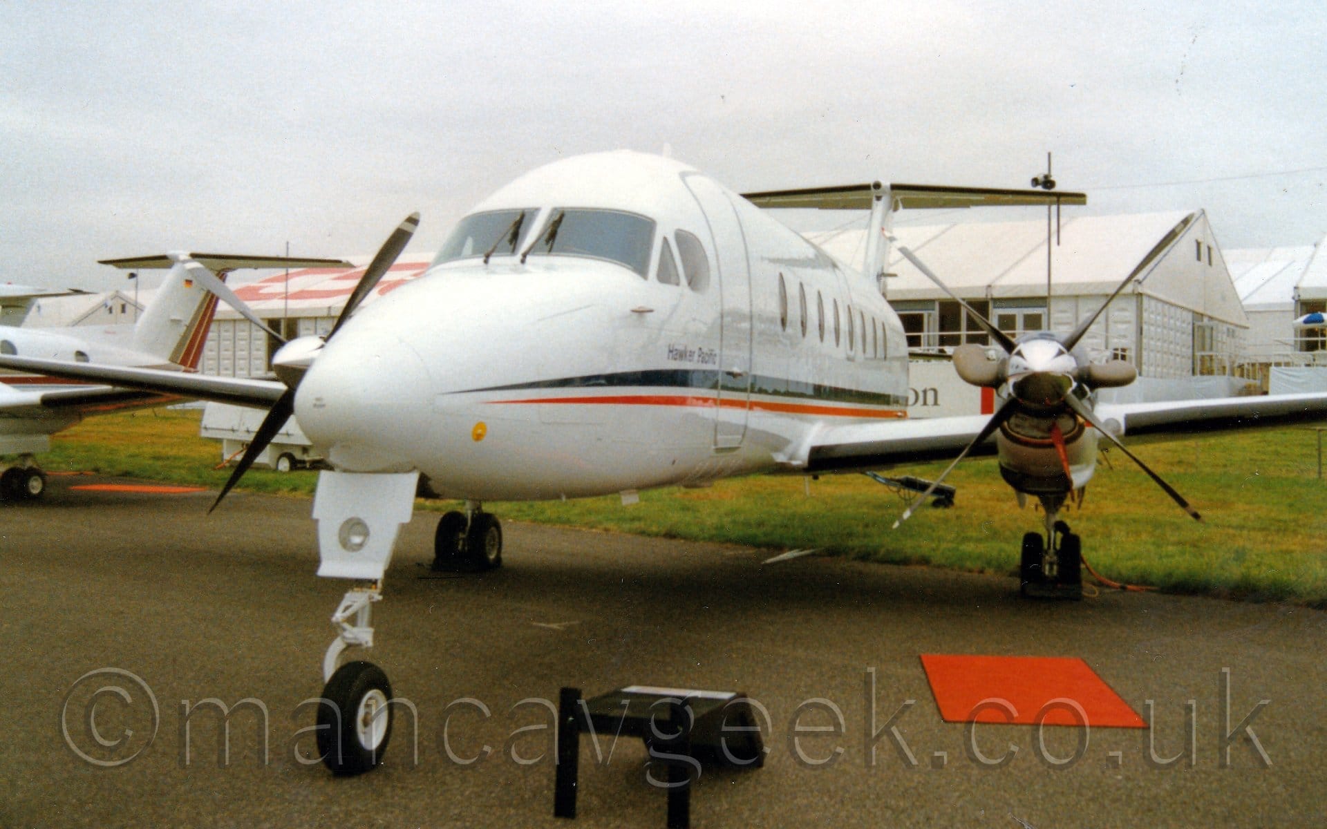 Forward view of a twin propellor-engined airliner parked on a taxiway facing slightly to the left of the camera. The plane is mostly white, with a black and orange stripe running along the body, and the words "Hawker Pacific" just below the cockpit side windows. There is a red mat on the floor, just in front of the plane's left engine, and a small black step beside the nose-wheel. Green grass in the background leads up to a series of white pre-fabricated buildings, under grey skies.