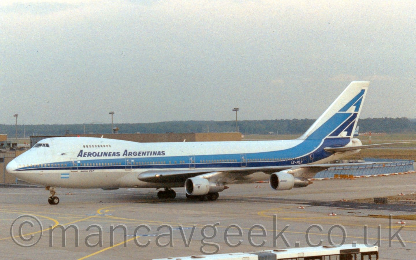 Side view of a very large, 4 engined jet airliner taxiing from right to left. The plane is largely white, with a light and dark blue strip running along the body, sweeping up into the tail, with a stylised pair of upper-case letter "A"'s. There are dark blue "Aerolieas Argentinas" titles on the upper forward fuselage, between the upper and lower deck windows. There is an Argentinian flag (light blue/white/light blue stripes) on the lower forward fuselage, and the white registration "LV-MLP" on the lower rear. Grey concrete taxiway marked with yellow lines fills the foreground, with a low brown building in the background, and trees in the distance, under flat grey sky.