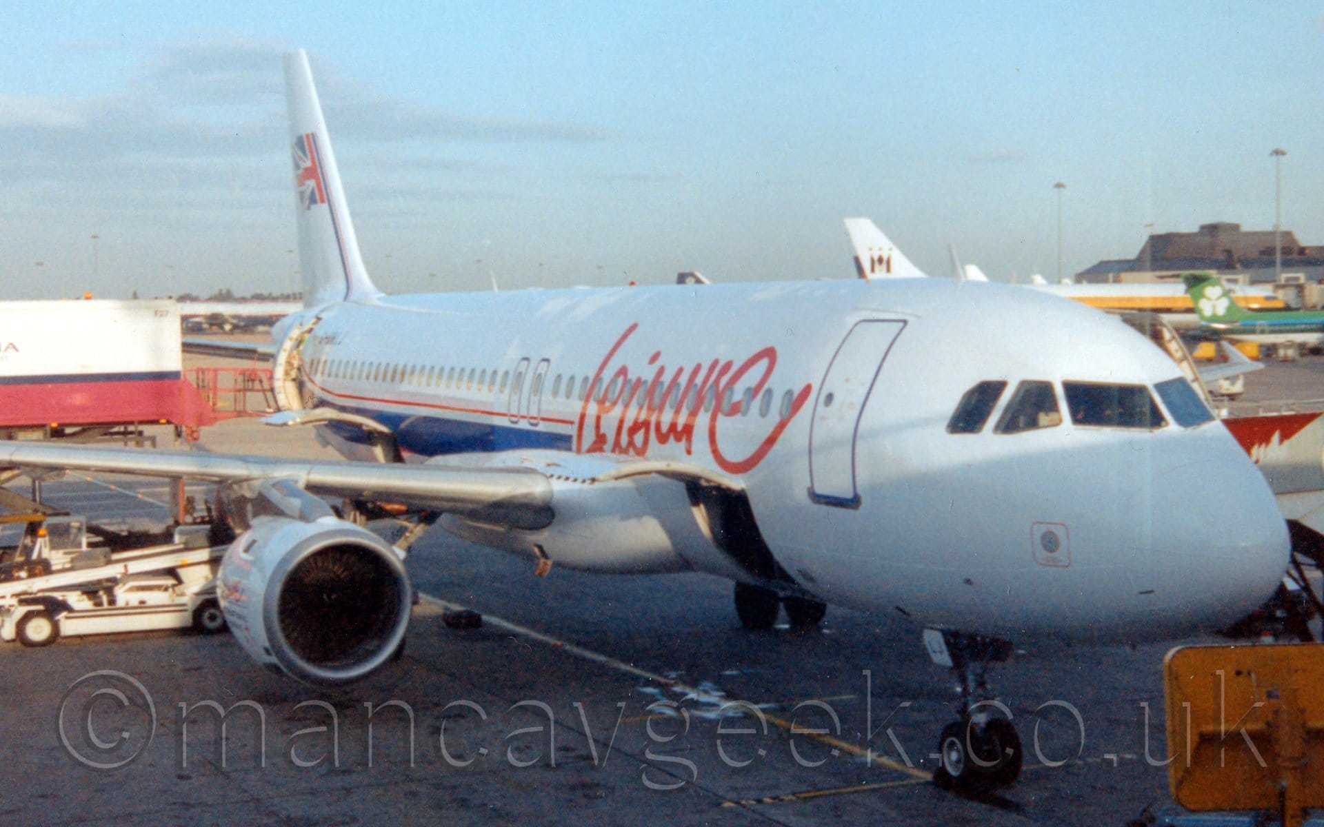 Forward side view of a twin engined jet airliner parked facing to the right. The plane is mostly white, with red "leisure" titles in a hand-written font on the forward fuselage, and a thin red pinstripe and a thick blue stripe running along the body under the cabin windows, before turning up into the tail where it becomes a lot thinner and has half of a British flag waving on the side. Cargo bay doors are hinged upwards both in front of and aft of the wings with a white baggage-loading belt raised to the rear door., and the rear cabin door is open, with a white and pink catering truck raised to gain access. Several other jet airliners can be seen parked by a terminal building in the background on the right of the frame. Pale blue sky with streaks of grey cloud fill the rest of the frame.