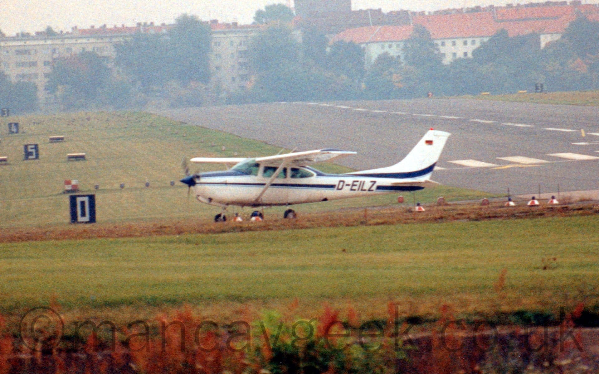 Side view of a high-winged, single engined light aircraft taxiing from right to left after landing on a black runway. The plane is white, with dark blue stripes running along the body and up into the bottom of the tail, with black registration "D-EILZ" on the rear fuselage. A wide, black runway stretches from the middle rear of the frame to the right edge, with green grass filling the foreground and the sides. There are black boards with white numbers on the left of the frame. White apartment buildings with red roofs are nestled in trees in the distance, under hazy, flat grey skies.