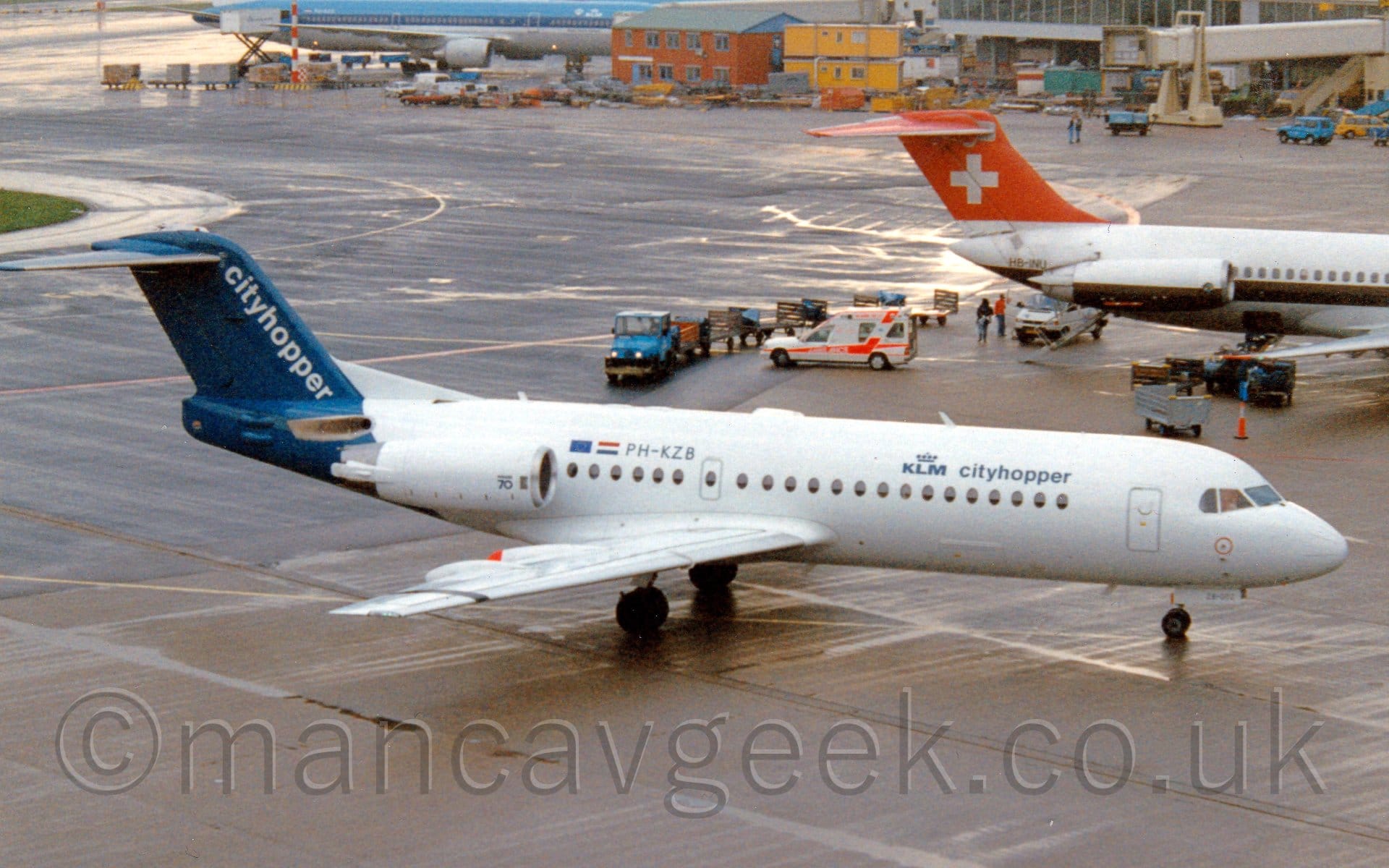 Side view of a twin engined jet airliner with the engines mounted on the sides of the rear fuselage, taxiing from left to right. The plane is largely white, with small, dark blue "KLM CityHopper" titles on the upper forward fuselage, with the registration "PH-KZB" on the upper rear, next to small EU and Dutch flags. The tail and rear fuselage is dark blue, with white diagonal "CityHopper" along the leading edge. Grey concrete apron fills the foreground and most of the background, as well as the background. The rear fuselage of a white jet airliner with a red and white tail is poking into frame on the right, with a blue and white jet airliner on the upper left., it's nose hidden behind a red brick building. A terminal building with a white air-bridge  is in the top right of the frame.