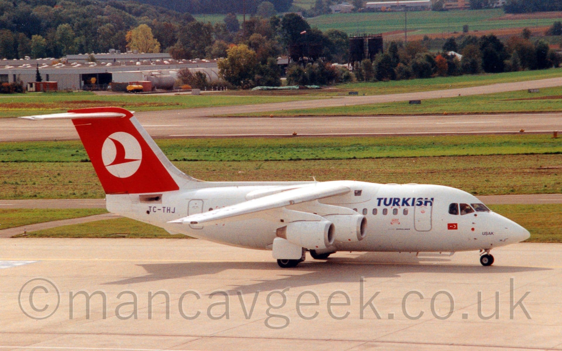 Side view of a high-winged, 4 engined jet airliner taxiing from left to right. The plane is almost entirely white, with blue "Turkish" titles on the upper forward fuselage, just in front of the wings. There is a small Turkish flag under the cockpit windows, and the blue name "Usak" on the nosecone, just above the nosewheel. The black registration "TC-THJ" is on the rear fuselage, under the tail. The tail is red, with a white circle containing a stylised flying bird. Grey concrete apron fills the foreground, with large grassed areas lining a grey runway and taxiway in the background, and buildings nestled in trees in the distance.