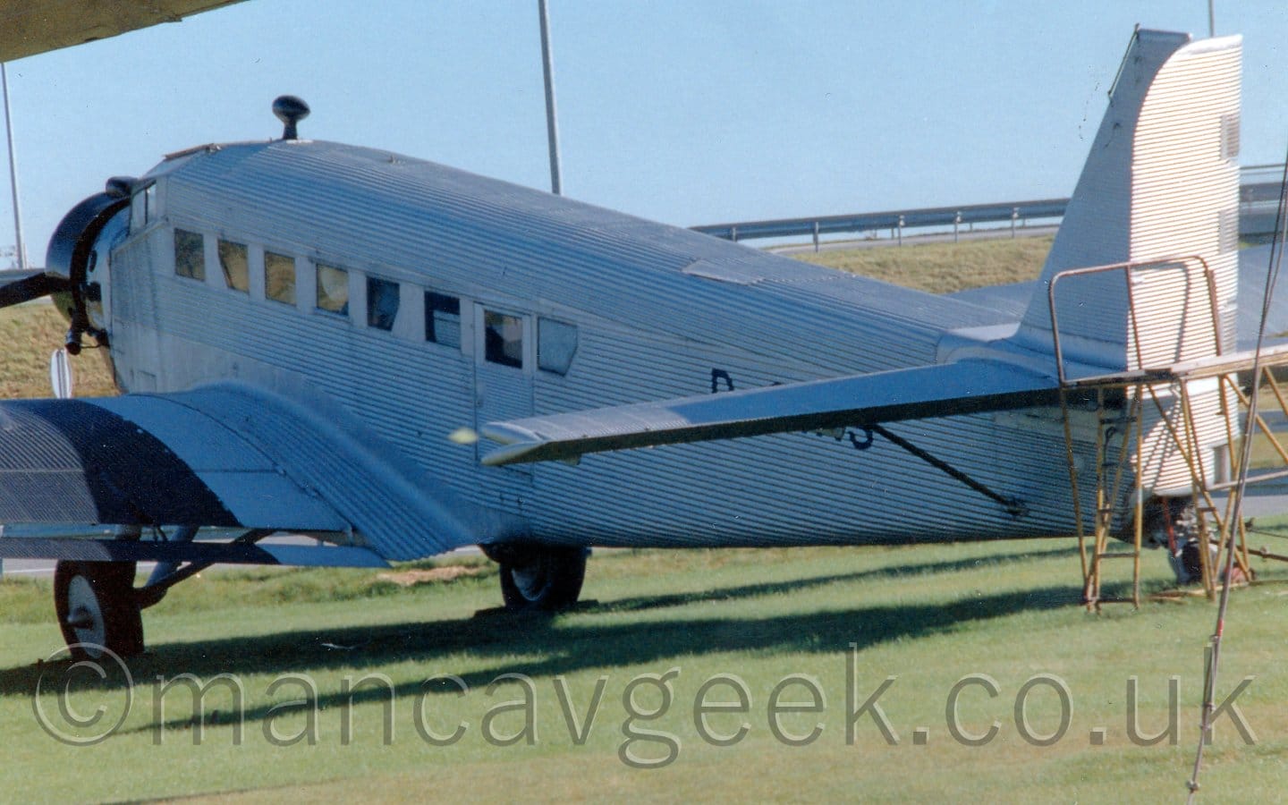 Side view of a 3 propellor-engined, World War 2-era airliner with a corrugated metal skin, parked on grass, facing to the left and away from the camera. The plane is in a metallic grey finish, with wide black stripes around the wings behind the engines, as well as a glossy black band around the engine in the motor. The black registration "D-CIAS" is on the rear fuselage, obscured by the horizontal stabiliser at the rear. Green grass fills the foreground, with a grassy bank with a metal fence along the top stretching across the background, under pale blue sky.
