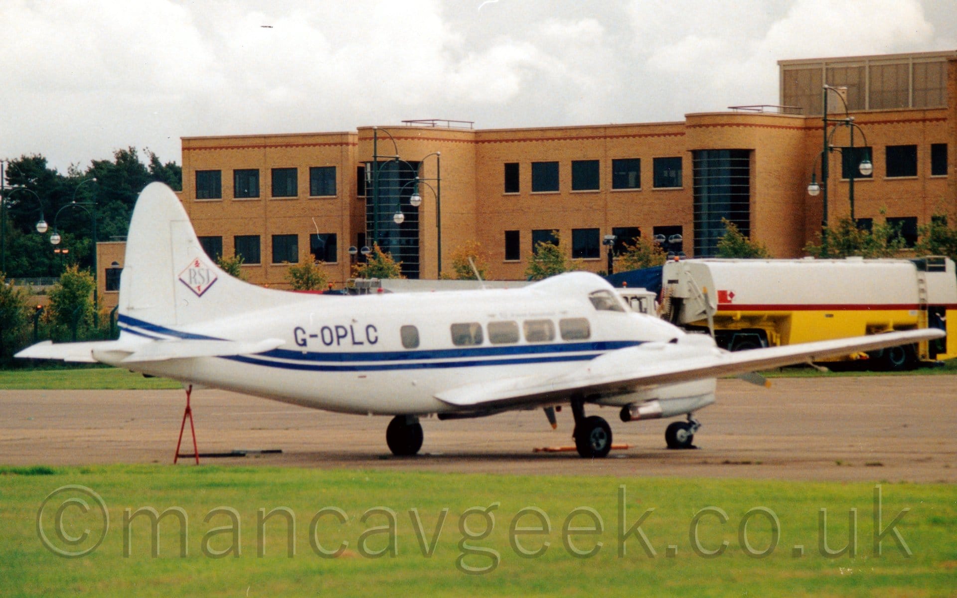 Side view of a small, white, twin propellor-engined airliner parked facing to the right and slightly away from the camera. The plane is mostly white, with a double blue stripe running along the body, and the blue registration "G-OPLC" on the upper rear fuselage. The cockpit is contained in a bubble higher than the passenger cabin, as can be seen with the difference between the cockpit and cabin windows. A triangular red frame is propping up the tail, resting on the ground. A white and red fuel tanker is parked in the background on the right of the frame, in front of a sandy-brown brick building with large, black windows. Cloudy grey sky fills the rest of the frame.