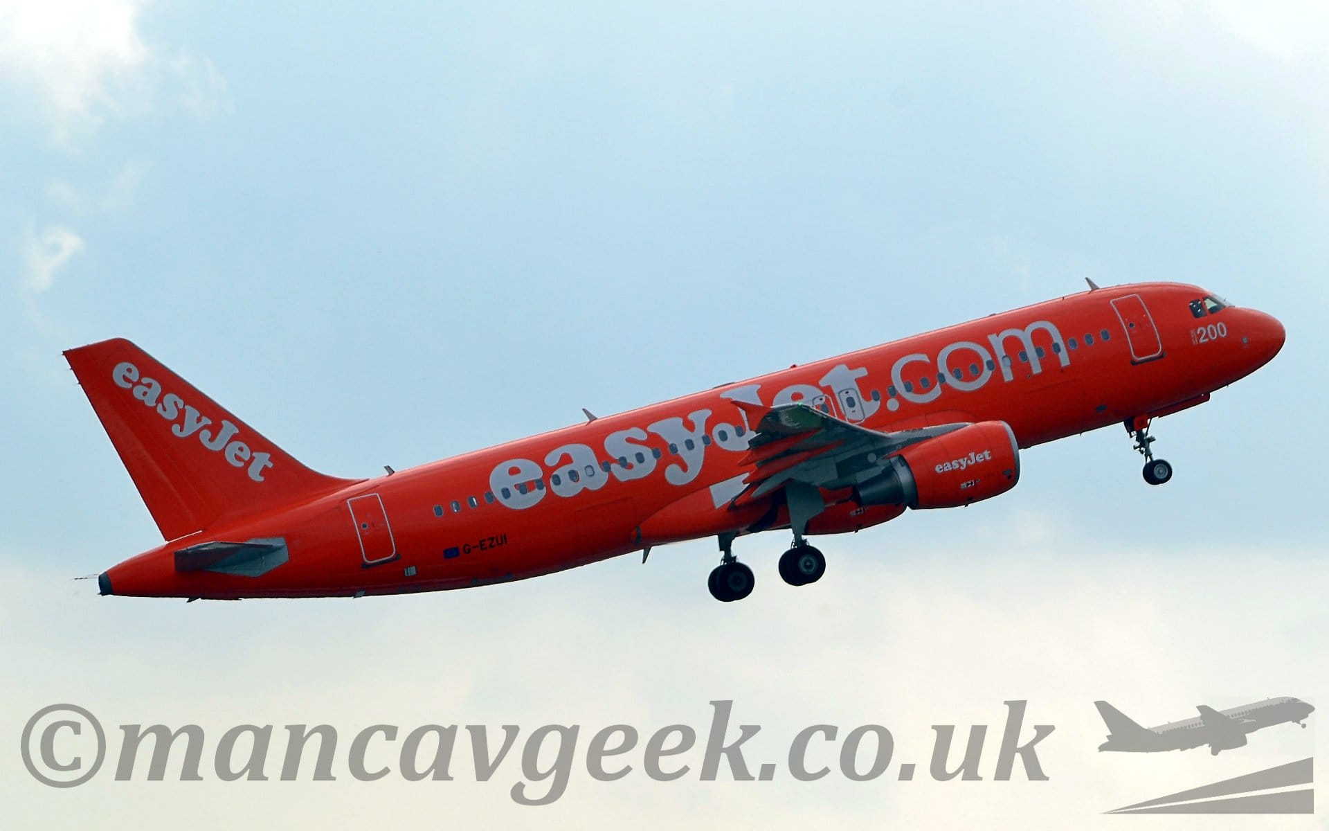 Side view of a bright orange, twin engined jet airliner flying from left to right at a low altitude, with undercarriage still lowered, flaps slightly extended from the rear of the wings, and the nose raised sharply, suggesting it has just taken off. There are large, white "EasyJet.com" titles written across the fuselage, with smaller "EasyJet" titles on the engine pods under the wings, as well as diagonally on the tail. There is a large white number "200" on the forward fuselage just under the cockpit windows, and the black registration "G-EZUI" on the lower rear fuselage. Pale blue sky with white patches of cloud fill the rest of the frame.