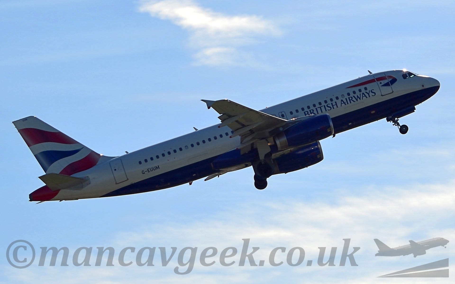 Side view of a twin engined jet airliner flying from left to right at a low altitude, with undercarriage starting to be tucked away, flaps extended from the rear of the wings, and the nose raised sharply, suggesting it has just taken off. The plane is mostly white, with blue "British Airways" titles on the lower forward fuselage, under the forward cabin windows, and a dark blue belly. The black registration "G-EUUM" is on the lower rear fuselage, under the rear cabin windows. The engine pods under the wings are also dark blue. The tail is white, with a pair of wavy red lines either side of a wavy blue triangle. Blue sky with smears of white cloud fill the rest of the frame.