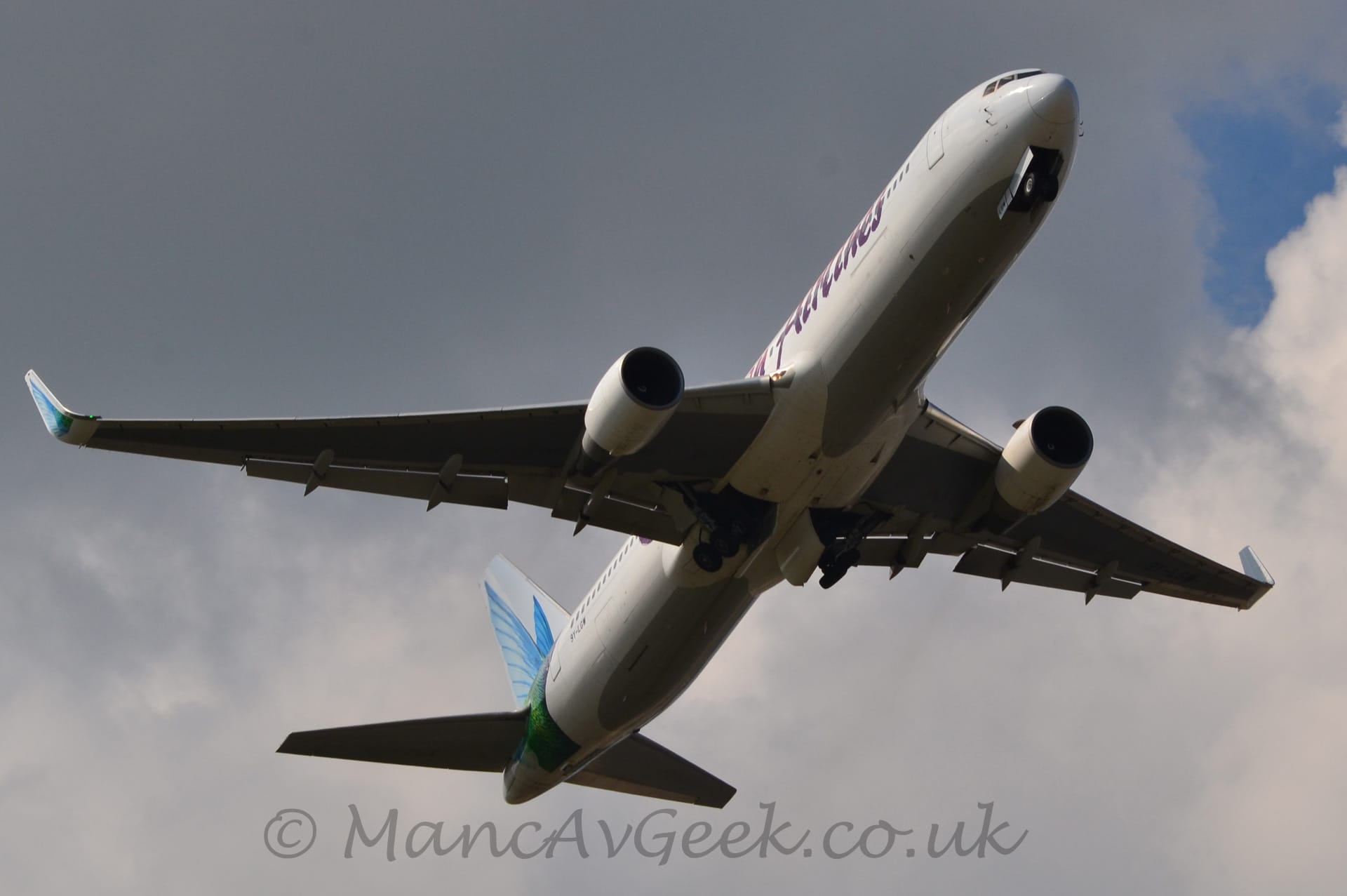 View from underneath of a twin-engined jet airliner flying from left to right at a low altitude, with it's undercarriage being tucked away, flaps extended from the rear of the wings, and the nose raised significantly, suggesting the plane has just taken off. The plane is mostly white with a grey belly, and large red "Airlines" text on the forward fuselage. There is a large image of a green bird with vivid blue outstretched wings on the rear fuselage and tail, as well on the up-turned wingtips. Dark grey clouds with patches of blue peeking through fill the rest of the frame.