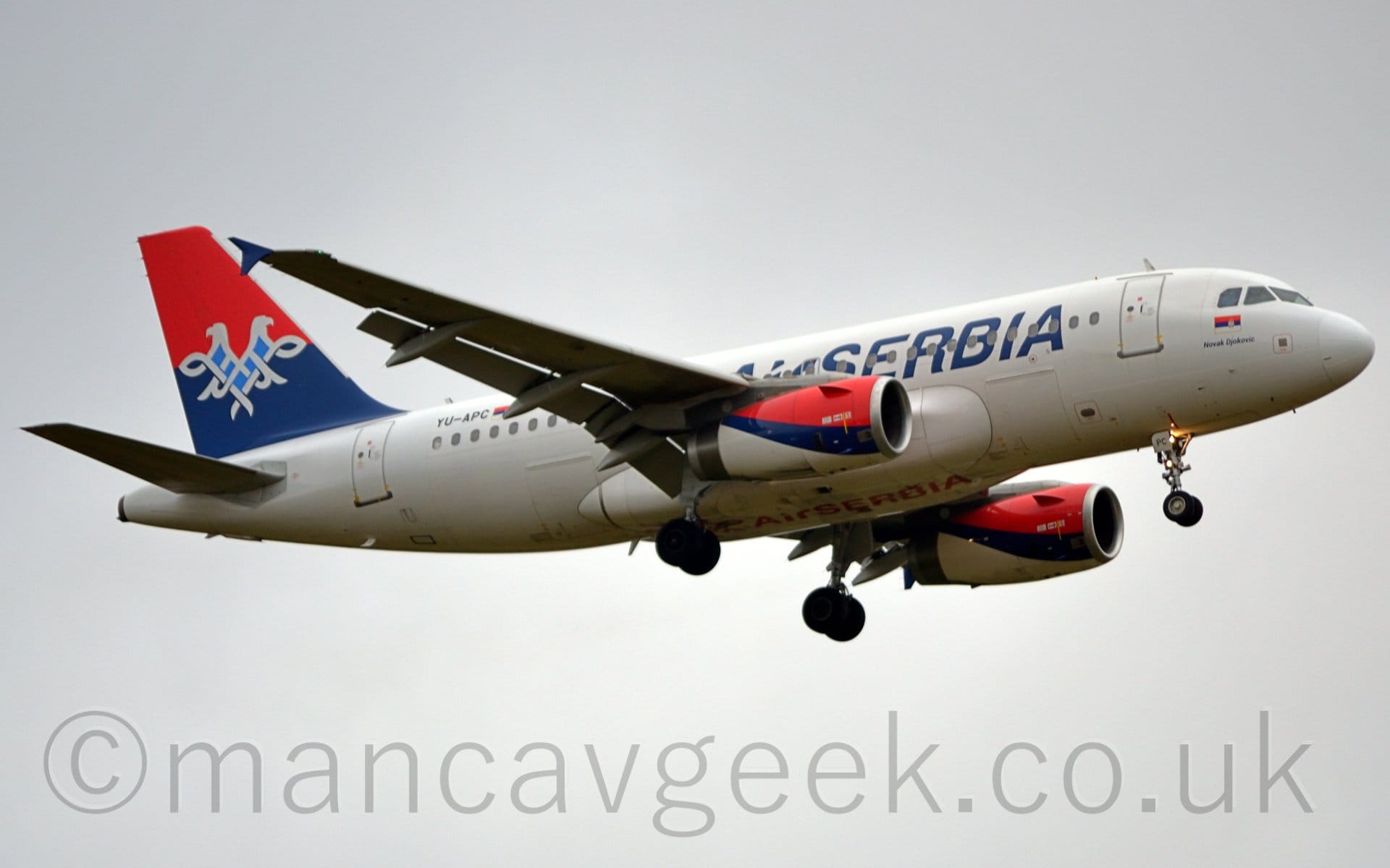 Low side view of a twin engined jet airliner flying from left to right at a very low altitude, with undercarriage lowered and flaps extended from the rear of the wings. The plane is mostly white, with blue, billboard-style "Air Serbia" titles on the forward fuselage, partially obscured by the wings, repeated in red under the body. The black registration "YU-APC" is on the upper rear fuselage next to a red, blue, and white Serbian flag. The top of the tail is red, with the base being blue, with a white scrollwork two-headed eagle with a pale blue chest overlaid on the 2 colours. The engine pods under the wings have a red top, white bottom, and diagonal blue tripe in the middle. Flat grey sky fills the background.