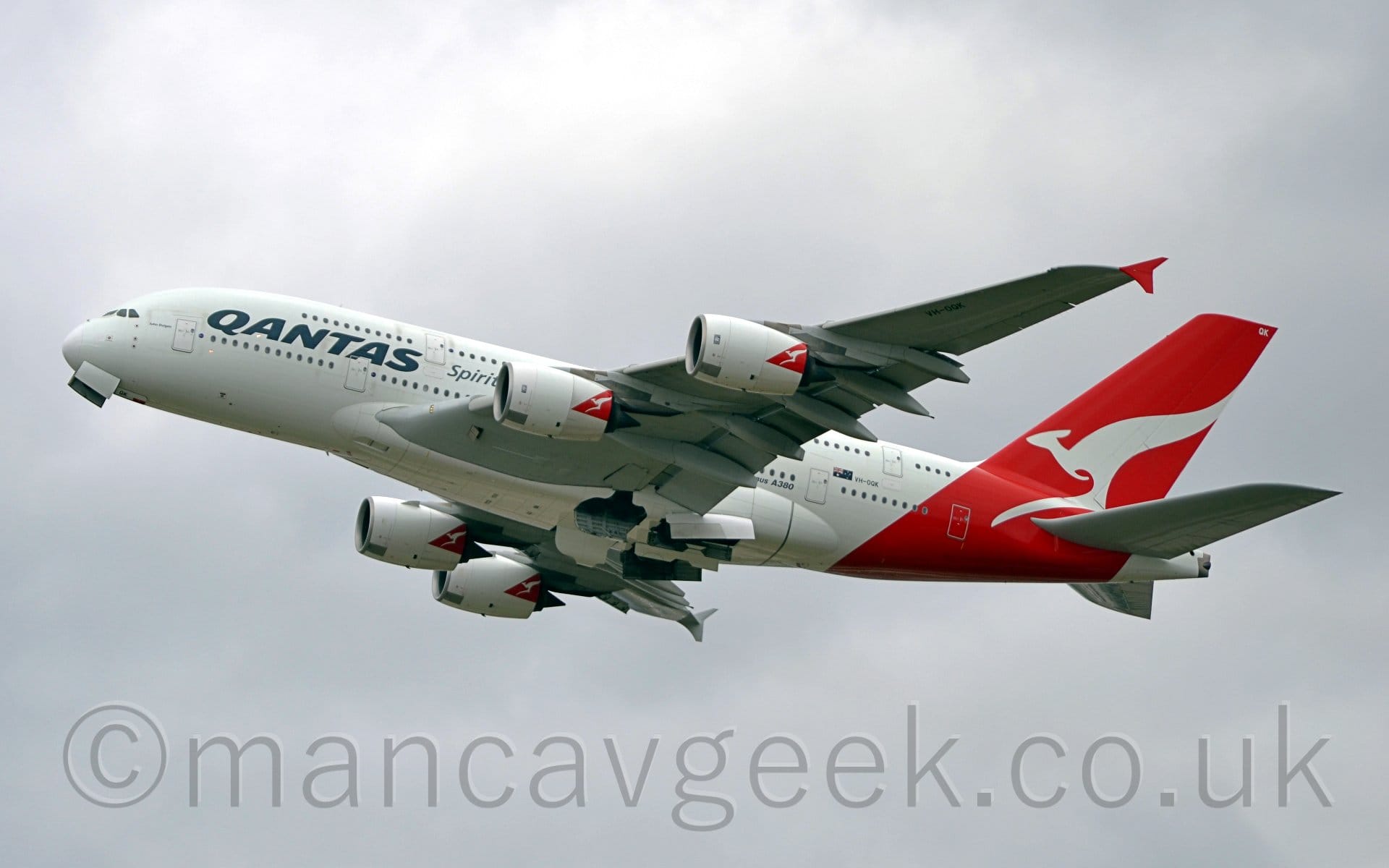 Low side view of a 4 engined jet airliner flying from right to left at a low altitude, with undercarriage being stowed away, and flaps extended from behind the wings, with the nose raised sharply, suggesting it has just taken off. The plane is largely white, with black "Qantas" titles on the upper forward fuselage, between the windows of the 2 passenger decks. The black registration "VH-OQK" is next to a small, Australian flag on the lower rear fuselage, near "Airbus A380" titles. The rear fuselage and tail is covered with a diagonal red band, which has the large silhouette of a white kangaroo in the middle, and the white letters "QK" right at the top. A red triangle with a small white kangaroo is on the upper rear of each of the 4 engines, mounted under the wings. The up- (and down-) turned wingtip fences are also bright red. Flat white clouds fill the background.