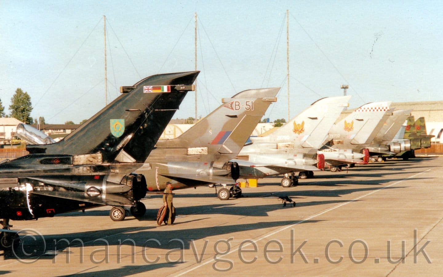 The tails of 10 fighter jets all parked side-by-side, ready and waiting for their turn to perform at an airshow, all facing to the left. The one on the left, nearest the camera, is in a gloss black livery, with the flags of the UK, Germany, and Italy at the top of the tail. Next is in a dark grey and brown camouflage, then 2 in light grey. 2 in dark grey follow, then 2 in pale blue, and finally 2 in green and brown camouflage. They are all casting strong shadows behind them on the ground, suggesting that light is coming from high on the left. 3 tall radio masts soar into the sky behind them, with a series of relatively low buildings behind, under pale blue sky.