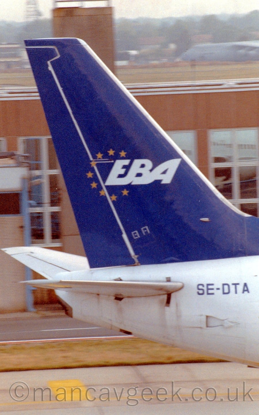 Vertically-oriented close-up of the blue tail of a white jet airliner taxiing from left to right. The black registration "SE-DTA" is on the upper rear of the fuselage, just below the tail. There is a circle of golden stars in the middle of the tail, wrapped around the white upper-case letters "EBA". A brown building with large glass windows stretches across the background, with a view across the airfield beyond that.
