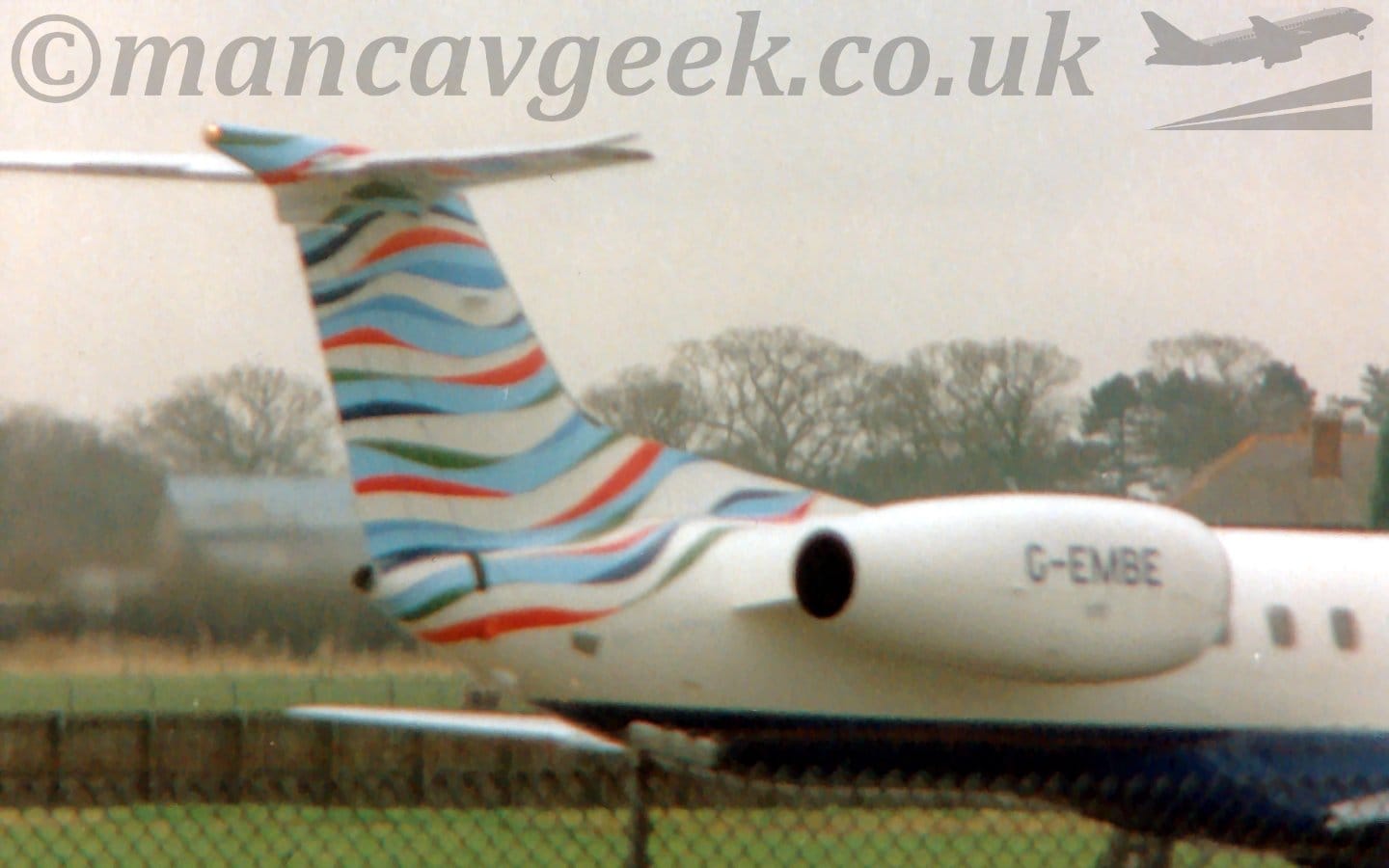 Slightly out-of-focus close-up of the rear fuselage and tail of a twin-engined jet airliner moving from left to right. The plane is mostly white, with a blue belly, almost random light and dark blue, red, and white curved stripes on the tail, and the black registration "G-EMBE" on the white engine pods mounted on the sides of the rear fuselage. Grass in the background leads up to trees in the distance, under hazy grey sky.