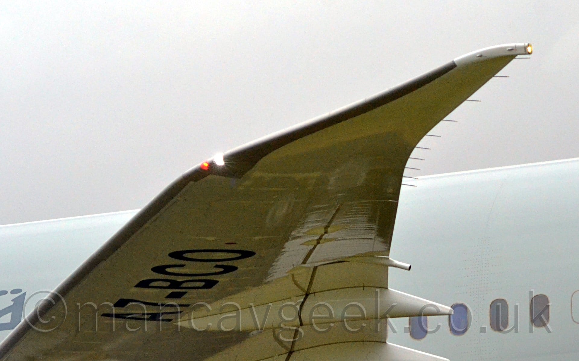 Close-up of the wingtip of a jet airliner taxiing from right to left. The plane has a grey body, with white wings, with the registration "A7-BCO" under the outer panels. A red and bright white light is shining in the corner of the wing just where the wingtip angles back. Flat grey sky fills the rest of the frame.