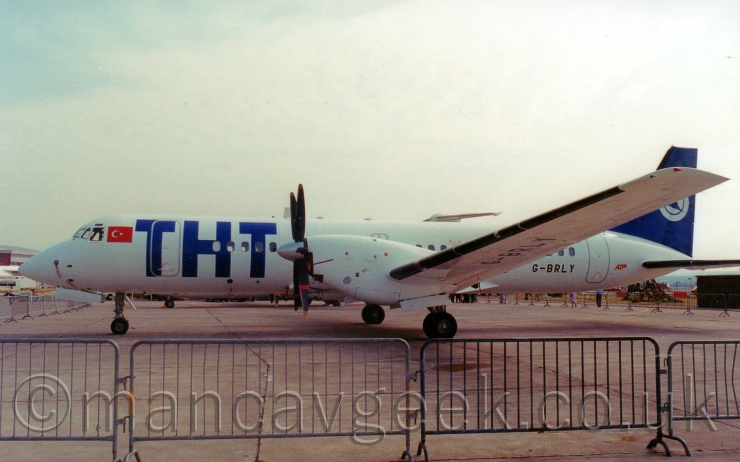 Side view of a twin propellor engined airliner parked facing to the left. The plane is mostly white, with large, blue, billboard-style "THT" titles on the forward fuselage, and the black registration "G-BRLY" on the lower rear fuselage, with the registration repeated under the left wing, outboard of the engine pod. The blue tail has a white circle in the middle, containing a stylised flying blue bird. A temporary metal fence stretches across the foreground, with the T-tail of another plane just visible over the top of this plane in the background, under flat grey skies.