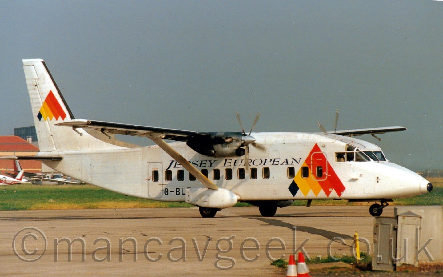 Side view of a high-winged, twin propellor-engined airliner taxiing from left to right. The plane is very squat and square, painted white over all with a large black, yellow, orange, and red-banded triangle with a feathered bottom edge on the forward fuselage, with black "Jersey European" titles over the passenger cabin windows under the wings, and the black registration "G-BLZT" on the lower rear fuselage, partially hidden by the sponsons carrying the main undercarriage. The multi-coloured triangle logo is repeated in a smaller form on the white tail. Stub wings jutting out from the bottom of the fuselage under the wing carry housings for the main gear to retract into, with a support strut rising diagonally from there towards the midpoint of the wing. Grey concrete apron fills the foreground, , with grass in the background, with parked planes in front of a red brick building on the left of the background, under blue-grey skies.