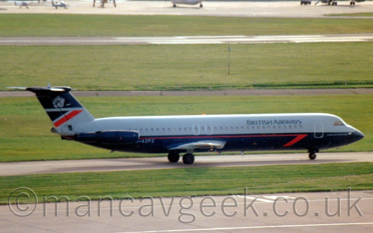 Side view of a twin engined jet airliner with the engines mounted on the sides of the rear fuselage, taxiing from left to right. The plane is dark grey on the top half of the body, and dark blue of the bottom half, with a red pinstripe just under the passenger cabin windows, ending in half an arrowhead under the forward fuselage. There are black "British Airways" titles on the upper forward fuselage, and the grey registration "G-AZPZ" just forward of the engine pods on the rear fuselage. The tail is dark blue at the top and grey at the bottom, with a dark blue triangle at the top rear of the grey part, and a diagonal red stripe below that. A grey heraldic crest is in the middle of the dark blue part at the top. Grey concrete apron and green grass fill the foreground, with more grass in the background leading up to a grey runway in the distance.