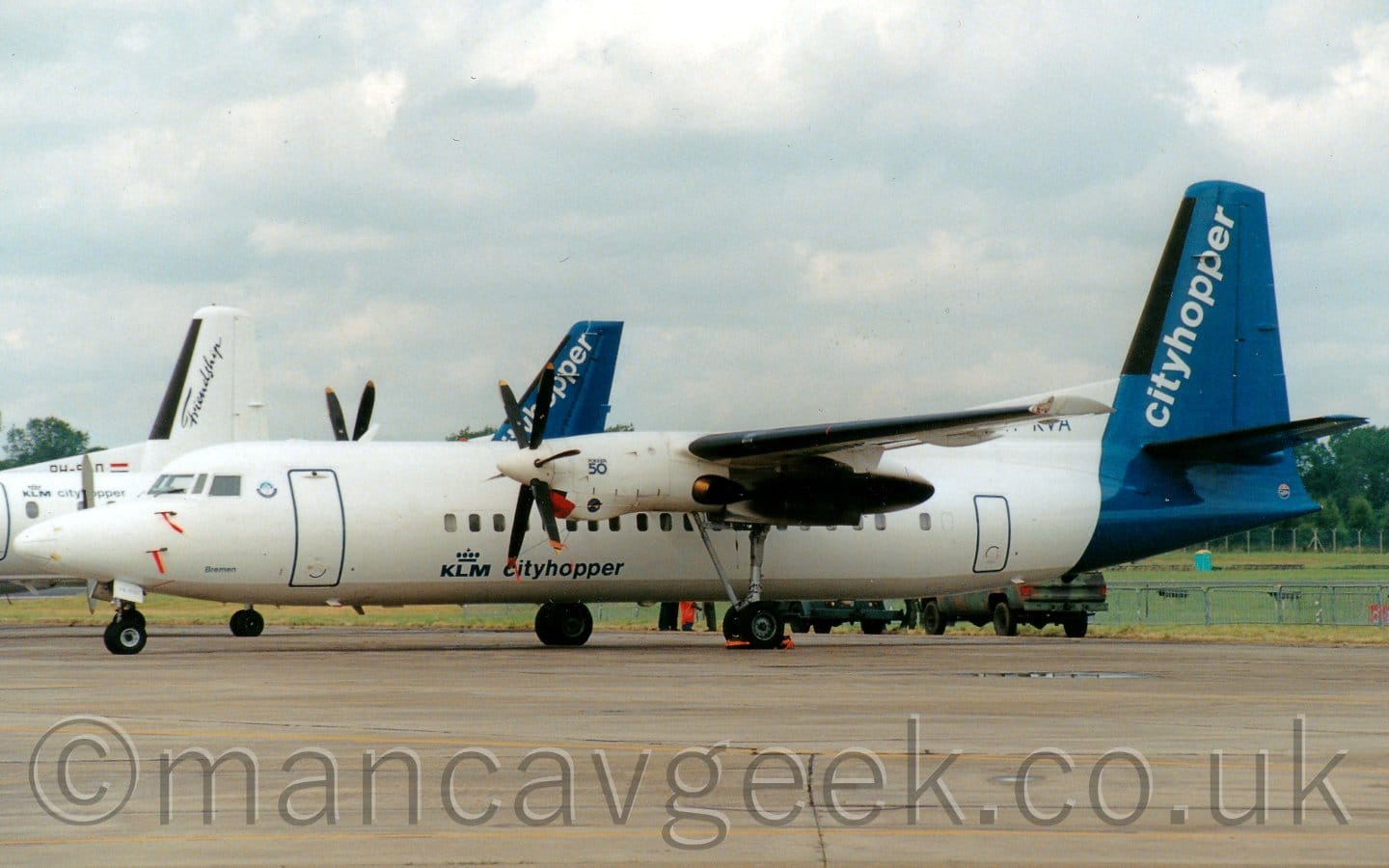 Side view of a high-winged, twin propellor-engined airliner parked facing to the left. The plane is mostly white, with a grey belly, and dark blue "KLM CityHopper" titles on the lower forward fuselage, with a stylised crown over the "KLM" part. The tail and rear fuselage are dark blue, with diagonal white "CityHopper" titles on the tail. Grey concrete apron fills the foreground, with grass in the background leading up to trees in the distance. The tail of another, smaller, CityHopper plane can be seen over the forward fuselage of this one, with another plane beyond that with the word "Friendship" on the white tail. Fluffy grey and white clouds fill the rest of the frame.