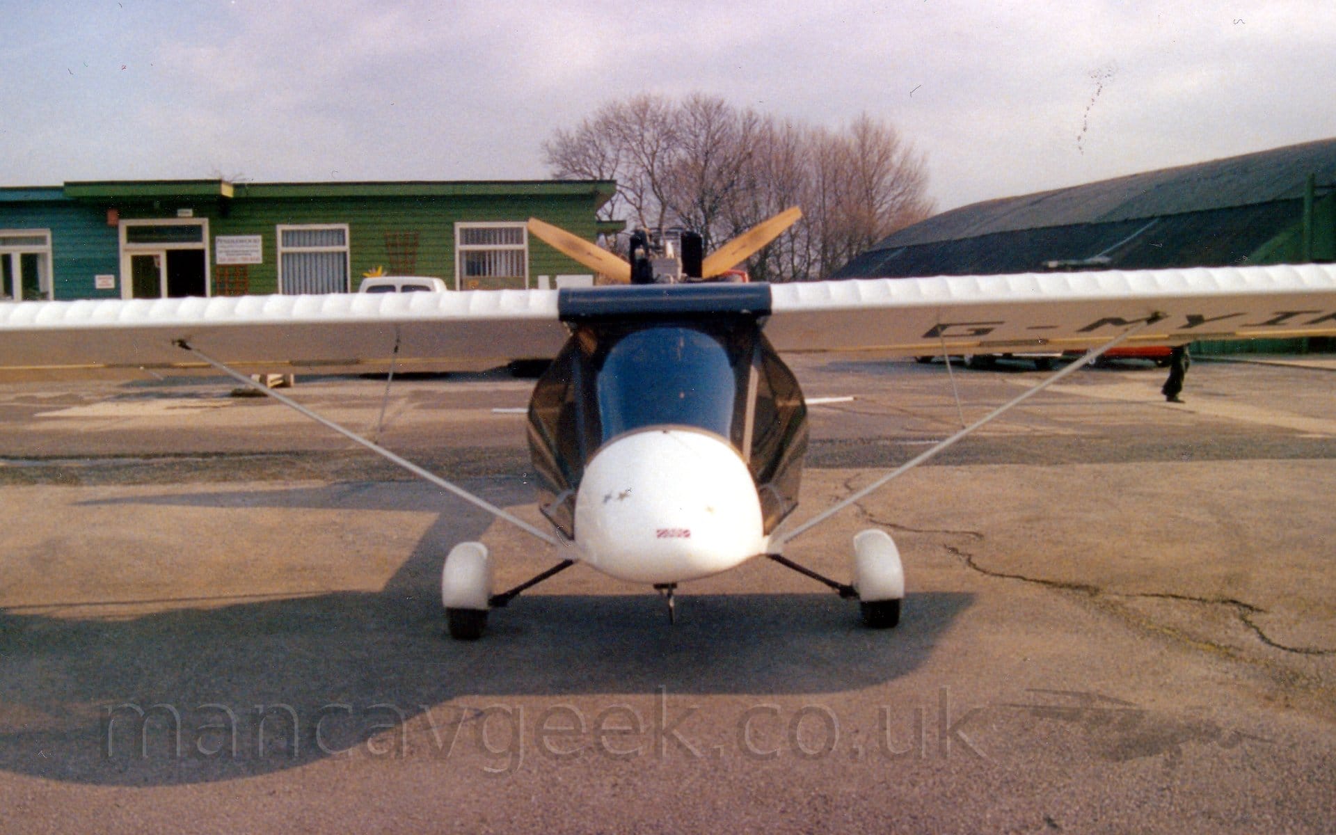 Head-on shot of a single-engined light aircraft, with the propellor mounted on the rear of the fuselage, facing backwards. A large, almost black perspex canopy gives great visibility forwards and to the sides for the occupants, although there is next to nothing ot the rear. The main wheels are on small struts coming out of the bottom of the fuselage, and are covered in white spats. A thin support strut comes out from the side of the fuselage by the undercarriage strut, rising diagonally to the mid-point of the rather thick, white wing. The black registration "G-MYIK" is visible under the left wing. The 3-bladed wooden prop is visible behind the cockpit, Grey tarmac apron fills the foreground, with a strong shadow being cast by the plane, sunlight coming from the right of the frame. A wooden, green building is in the background on the left, with a green hangar with a black domed roof on the right, and trees beyond in the middle. Bright but hazy sky fills the rest of the frame.