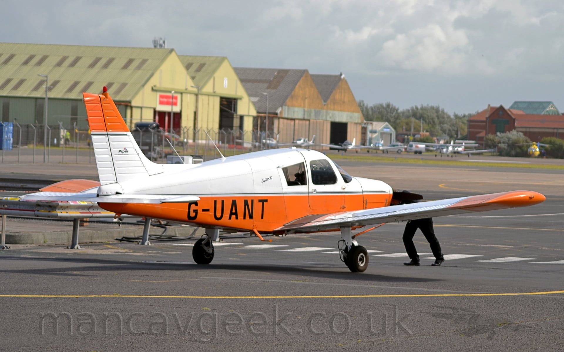 Side view of  a single engined light aircraft facing away from the camera and slightly to the right, being pushed backwards by a person in black holding on to the propellor at the front. The plane is white on top and orange underneath, with the black registration "G-UANT" on the lower rear fuselage. The tail has the colours reversed, white at the bottom, and orange at the top. The wings are also white, with the outer panels being orange. Black tarmac apron fills the foreground, with more apron in the background, with some more light aircraft parked in front of hangars in the distance. Cludy white and grey sky with the occasional hint of blue fills the rest of the frame.
