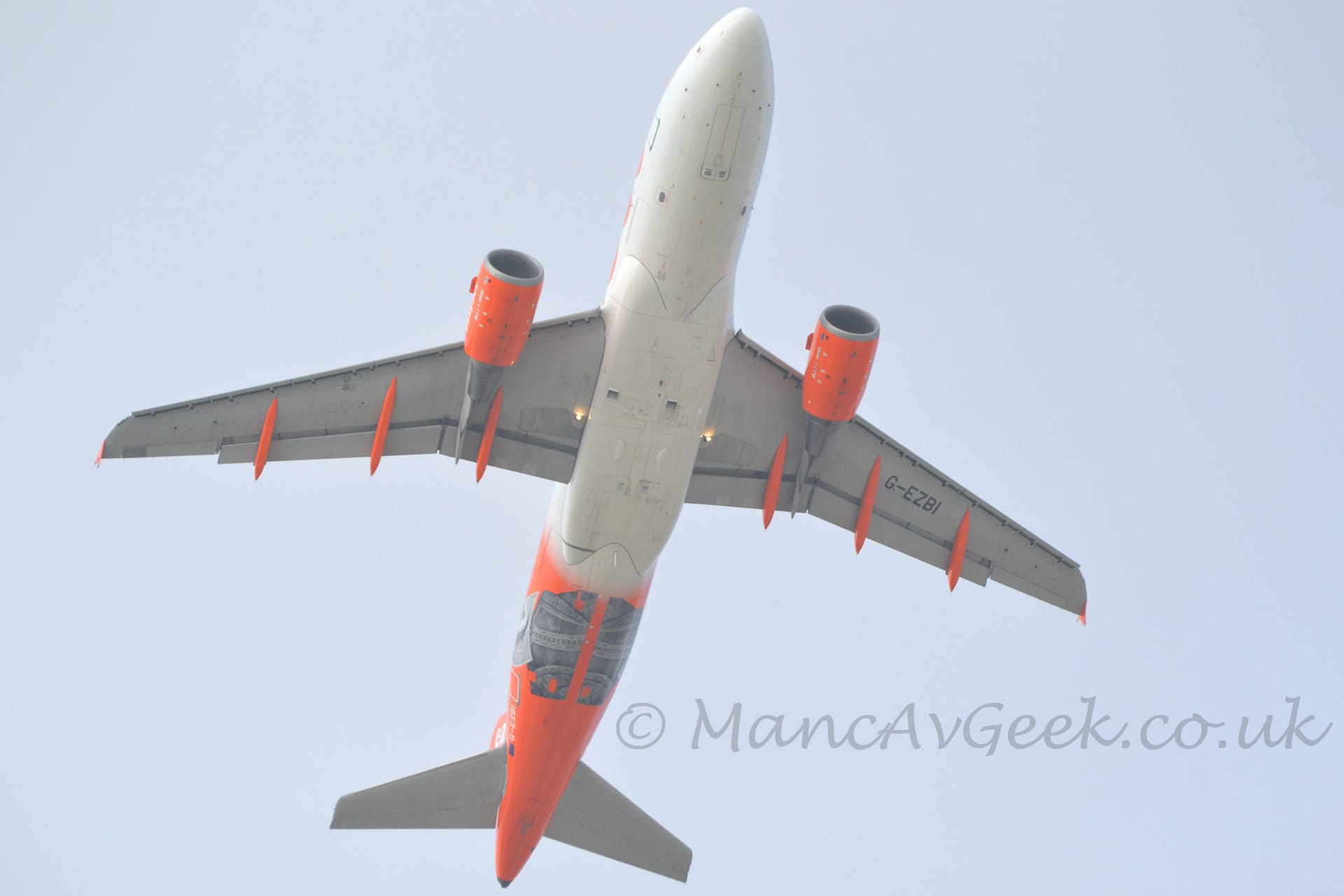 View from below o a twin engined jet airliner flying pretty much directly overhead at a very low altitude, with undercarriage tucked away, flaps extended from the rear of the wing, and the nose raised, suggesting it has just taken off. The plane is largely white, with an orange rear fuselage overlaid with the grey head and shoulders of a historical figure. The registration "G-EZBI" is on the lower rear fuselage in white, and under the left wing in black. The engine pods under the wings, as well as the flap track covers, are also bright orange. Pale grey-blue sky fills the background.