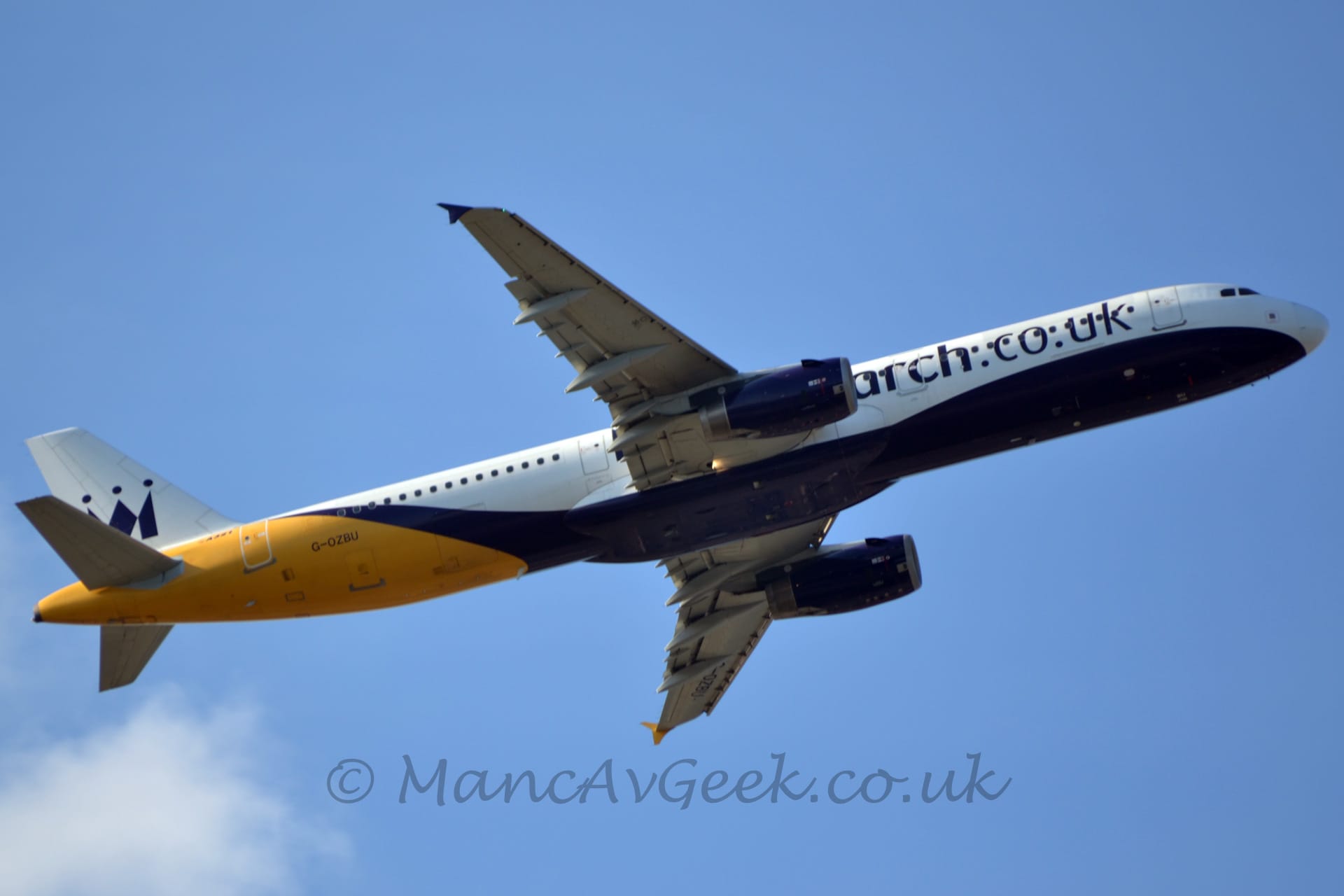 Side view of a twin engined jet airliner flying from left to right at a low altitude, with undercarriage stowed, flaps extended behind the wings, and the nose raised sharply, suggesting it has just taken off. The plane is mostly white, with a deep purple belly the sweeps up toward the top of the rear fuselage, bright yellow rear fuselage, and deep purple web address "monarch.co.uk" on the forward fuselage, covering the forward cabin windows. The black registration "G-OZBU" is on the lower rear fuselage, with a stylised black crown that more than resembles an upper case letter "M" on the white tail. Blue sky fills the rest of the frame, with a fluff of white cloud in the bottom left corner.