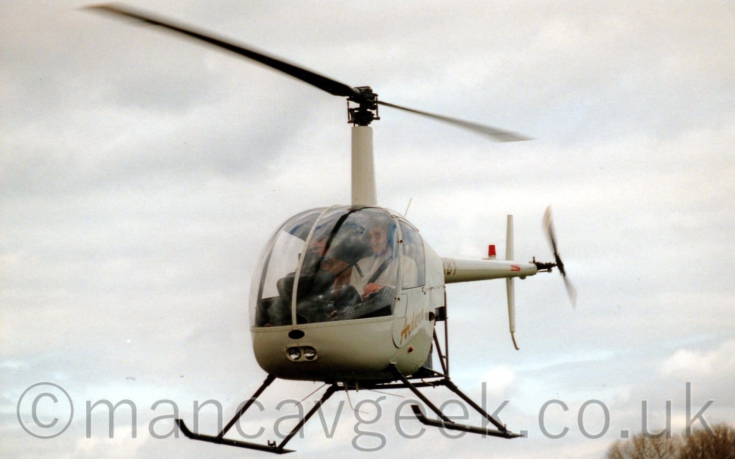 A small, 2 seat helicopter hovering a feet above the ground, facing slightly to the left of the camera, with both occupants clearly visible through the large cockpit windows. The 'copter is almost entirely white, with golden, seemingly handwritten "Ardern" titles on the cockpit door, and the golden registration "G-BRBY" on the thin tail boom, the first couple of letters obscured by the rear of the fuselage Some bare trees are visible in the bottom right of the frame, with fluffy grey and white clouds filling the rest of the frame.