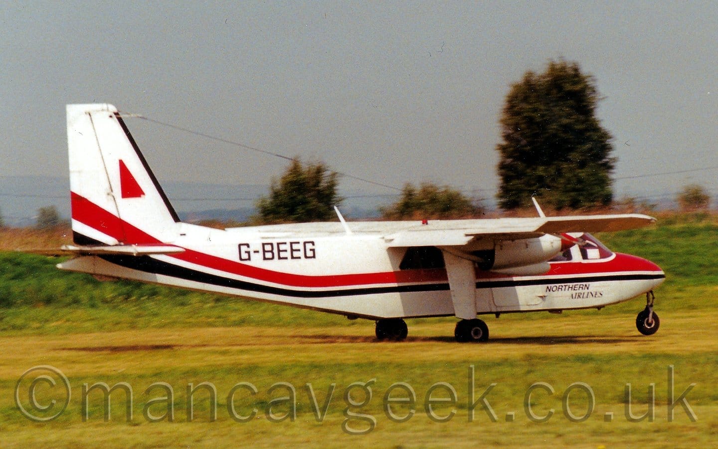 Side view of a high-winged, twin propellor-engined airliner moving at speed from left to right, with flaps extended from the rear of the wings, and the nose-wheel slightly off the ground, suggesting it is taking off. The plane is largely white, with a red and black stripe running along the body, sweeping up into the lower reaches of the tail. There is a red triangle towards the front of the tail. The black registration "G-BEEG" is on the upper rear fuselage, and smaller, black "Northern Airlines" titles under the cockpit. Green grass fills the foreground, and much of the background, with trees in the distance and grey skies over everything else.