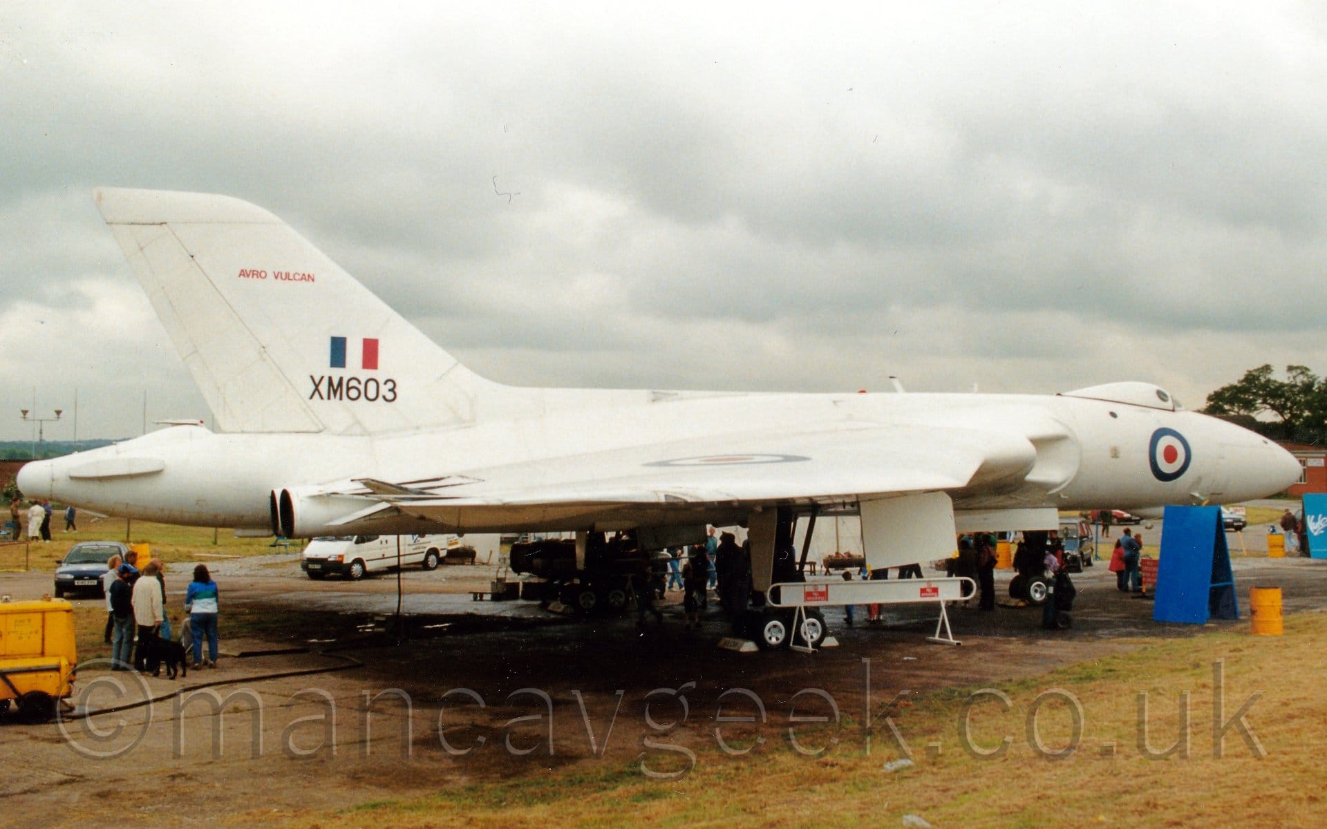 Side view of a delta-winged, 4 engined military bomber aircraft parked facing to the right. There is a blue, white, and red roundel on the nose, under the bump that is the cockpit, with another roundel on top of the right wing. The black serial "XM603" is on the power part of the tail, under a blue, white, and red fin flash, with red "Avro Vulcan" titles towards the top of the tail. There are people clustered under the belly of the plane, with another group of people under the tail, looking up at the engine exhausts, coming out of the rear of the wings, next to the rear fuselage. Green grass fills the foreground, with fluffy grey clouds filling the rest of the frame.
