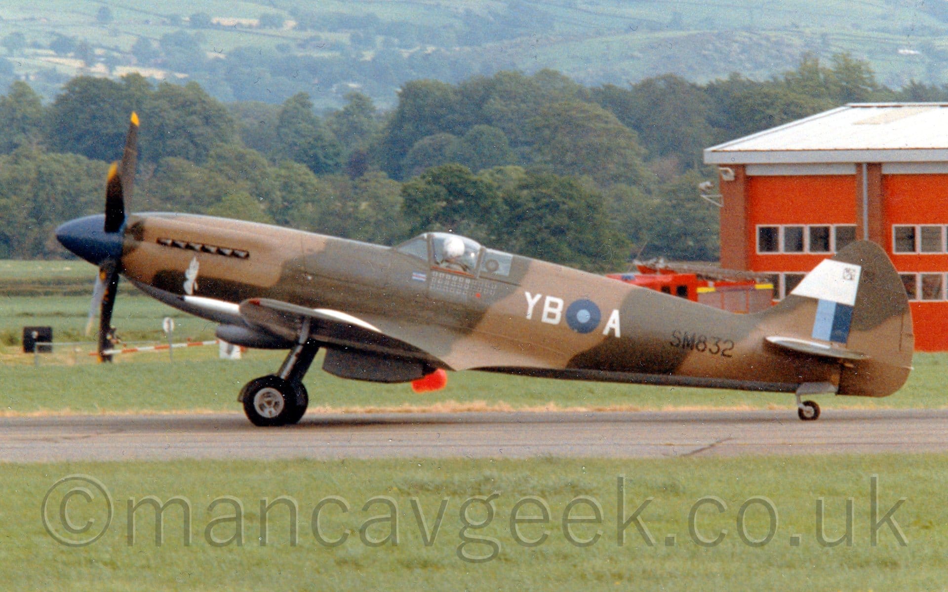 Side view of a single seat, piston engined fighter aircraft taxiing from right to left along a black runway. The plane is covered in a light and dark brown camouflage scheme, with a grey belly. There is a dark and light blue roundel on the rear fuselage, with the white letters "YB" at the front, and "A" behind, and the black serial "SM832" just in front of the tail The tail carries a light and dark blue fin flash under an all-white section. Green grass fills the foreground, with more grass in the background leading up to a brown brick building with large red doors, and a fire engine parked outside, on the right, and fields and trees on the left, with gently rolling hills in the distance.
