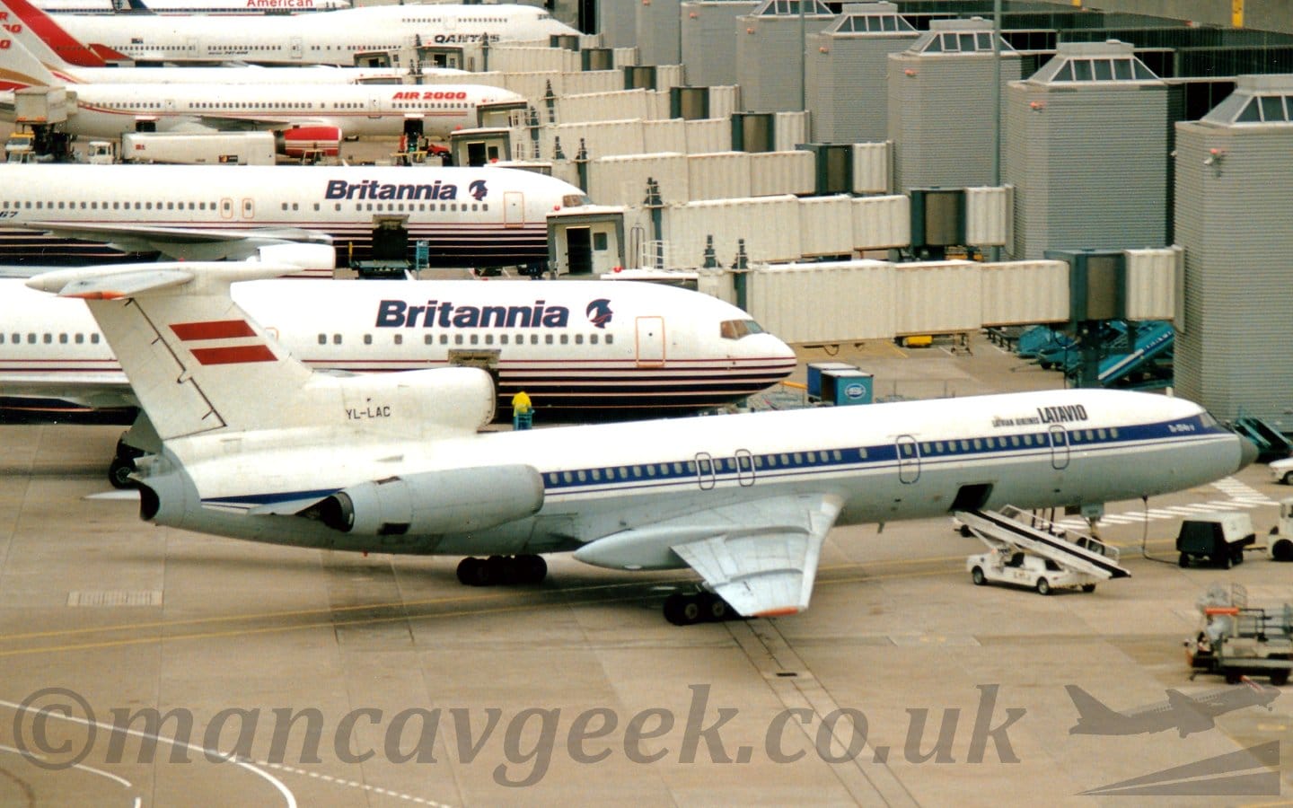 Side view of a 3 engined jet airliner parked by a terminal, facing to the right. The plane has a white upper fuselage, with grey on the lower section, and a blue stripe running along the body, covering the passenger cabin windows. There are black "LatAvio" titles on the upper forward fuselage, with smaller white text "Tu154B-2" on the blue stripe just aft of the cockpit. The black registration "YL-LAC" is on the centre engine intake, just in front of the tail, which itself has a large Latvian flag (2 thick red horizontal stripes separated by a thin white stripe) in the middle. The other engines, one on each side mounted on the rear fuselage, are grey. A baggage compartment hatch is open in the lower fuselage, between the forward- and mid-cabin doors, with a mobile baggage-loading belt raised towards it. Grey concrete apron fills the foreground, with a large terminal building on the right of the frame, and several mostly white airliners parked nose in, with white airbridges attached to the far sides.