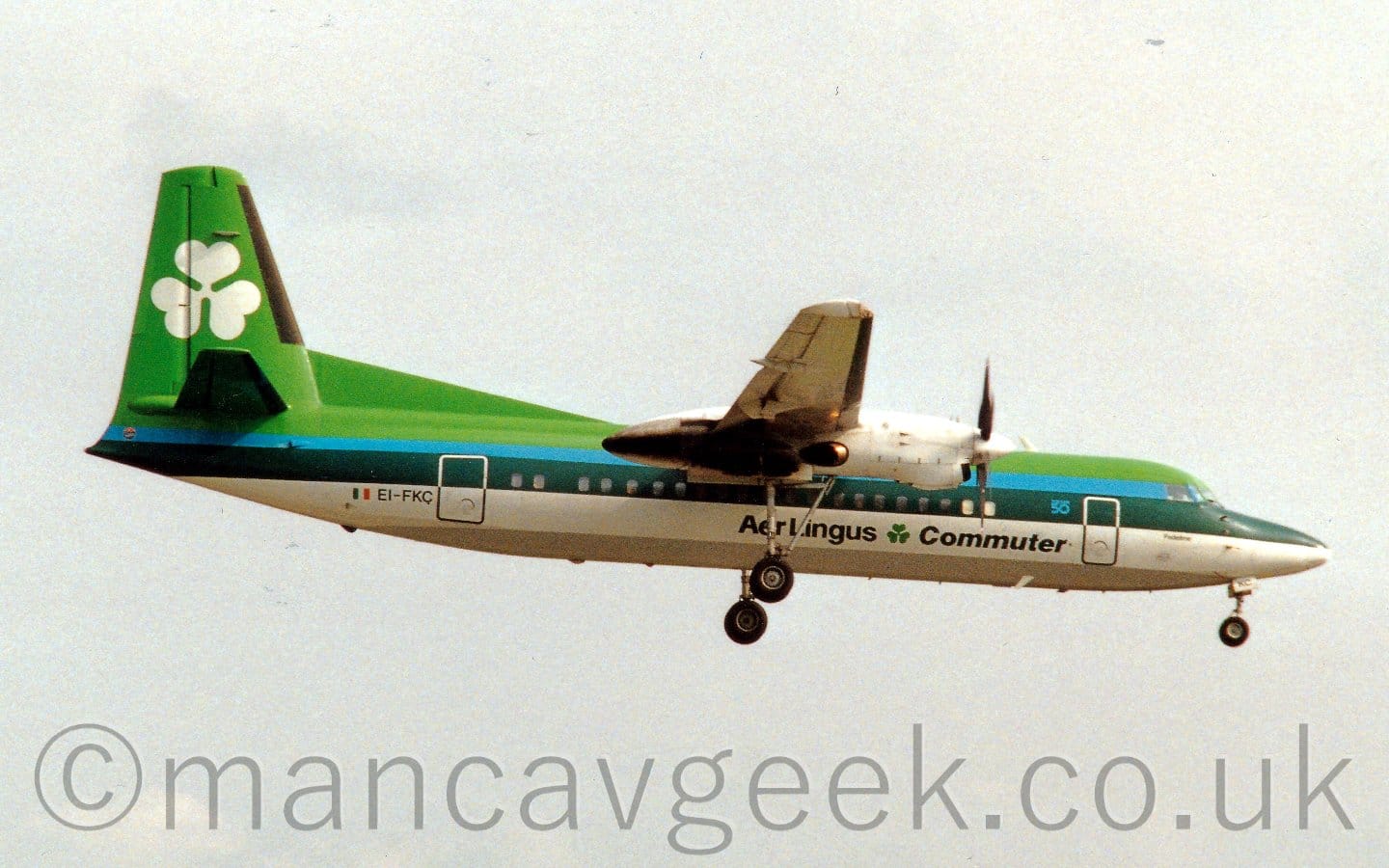 Side view of a high-winged, twin propellor-engined airliner flying from left to right at a low altitude, with undercarriage extended from the engine pods on the wings, and flaps extended from behind the wings, suggesting it is about to land. The plane has a bright green upper fuselage, with a pale blue and thick dark green stripe running along the body, a thick white stripe below that, and a grey belly. There are black "Aer Lingus Commuter" titles on the lower forward fuselage, with a green shamrock in the middle. A large white shamrock fills the green tail. The black registration "EI-FKC" is on the lower rear fuselage, next to a small Irish flag. Flat grey sky fills the rest of the frame.