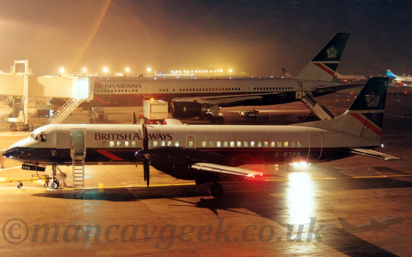 Night-time side view of a twin propellor-engined airliner parked facing to the left. The plane has a dark blue lower fuselage, with the upper fuselage being dark grey, with black "British Airways" titles on the upper forward fuselage. The grey registration "G-BTPD" is on the lower rear fuselage The forward door is open, with a built-in set of airstairs deployed. A larger, twin engined jet airliner in a similar livery is parked on the next gate, with an air bridge attached to the forward door. Bright lights from airport lighting stands, vehicles, and other planes illuminate the scene, blowing out some parts of the image with the long exposure required to bring out details.
