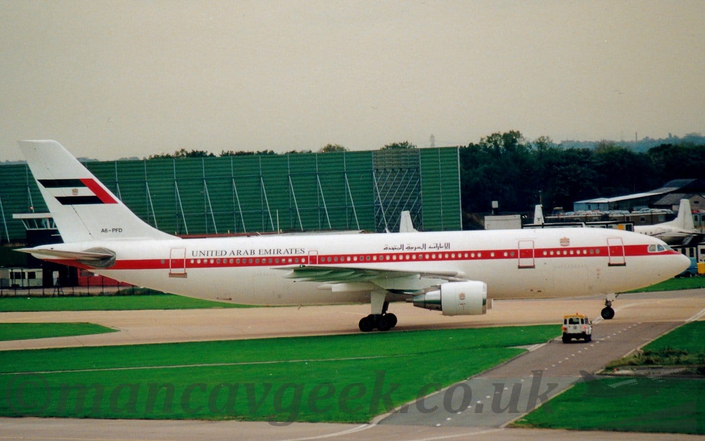 Side view of a twin engined jet airliner taxiing from left to right. The plane is largely white, with a red stripe running along the body, covering the passenger cabin windows. There are black "United Arab Emirates" titles on the upper fuselage, in an Arabic script above the wings, and in English at the rear. The black registration "A6-PFD" is on the base of the tail, which has a large United Arab Emirates flag in the middle. Lush green grass fills the foreground, with a black roadway splitting it on the grass, with a white vehicle with a yellow roof waiting for the plane to taxi past. A large green metal frame dominates the background on the left, with the tail of a smaller white airliner nestled amongst small buildings, with trees behind. Bright but hazy sky fills the rest of the frame.
