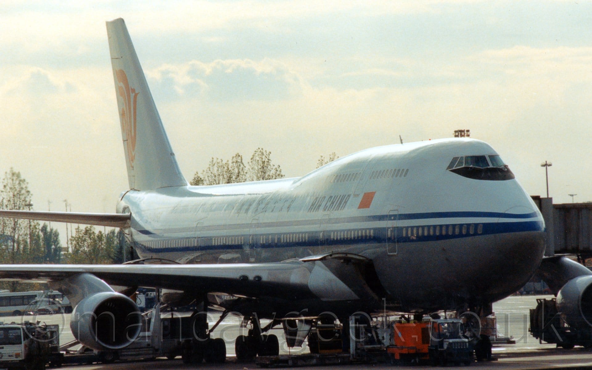 Side view of a very large, 4 engined jet airliner parked facing to the right, with a grey airbridge attached to the forward door on the opposite side, and various airport vehicles scattered around in front of it. The plane is white on top, with a grey belly, and a thick and a thin blue stripe running along the body, covering the lower deck windows.. There are black "Air China" titles next to a Chinese flag on the upper fuselage, between the upper and lower passenger deck windows. Scattered trees line the skyline, with bright but hazy sky with blue patches filling the rest of the frame.