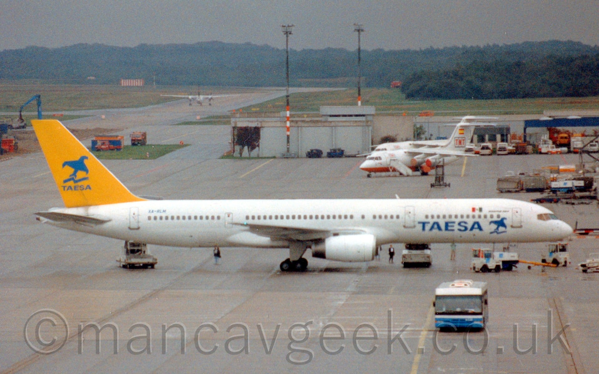 Side view of a twin engined jet airliner parked facing to the right, surrounded by several white airport vehicles. The plane is almost entirely white, with blue "TAESA" titles on the lower rear fuselage, next to a stylised image of a flying blue bird. The blue registration "XA-RLM" is on the upper rear fuselage, just ahead of the tail. The tail is bright yellow, with a larger version of the blue flying bird logo, over smaller "Taesa" titles. Grey concrete apron fills the foreground and most of the background, with a pair of white and orange, high-winged, 4 engined jet airliners on the right of the frame, with trees in the distance, uner dreary grey sky.