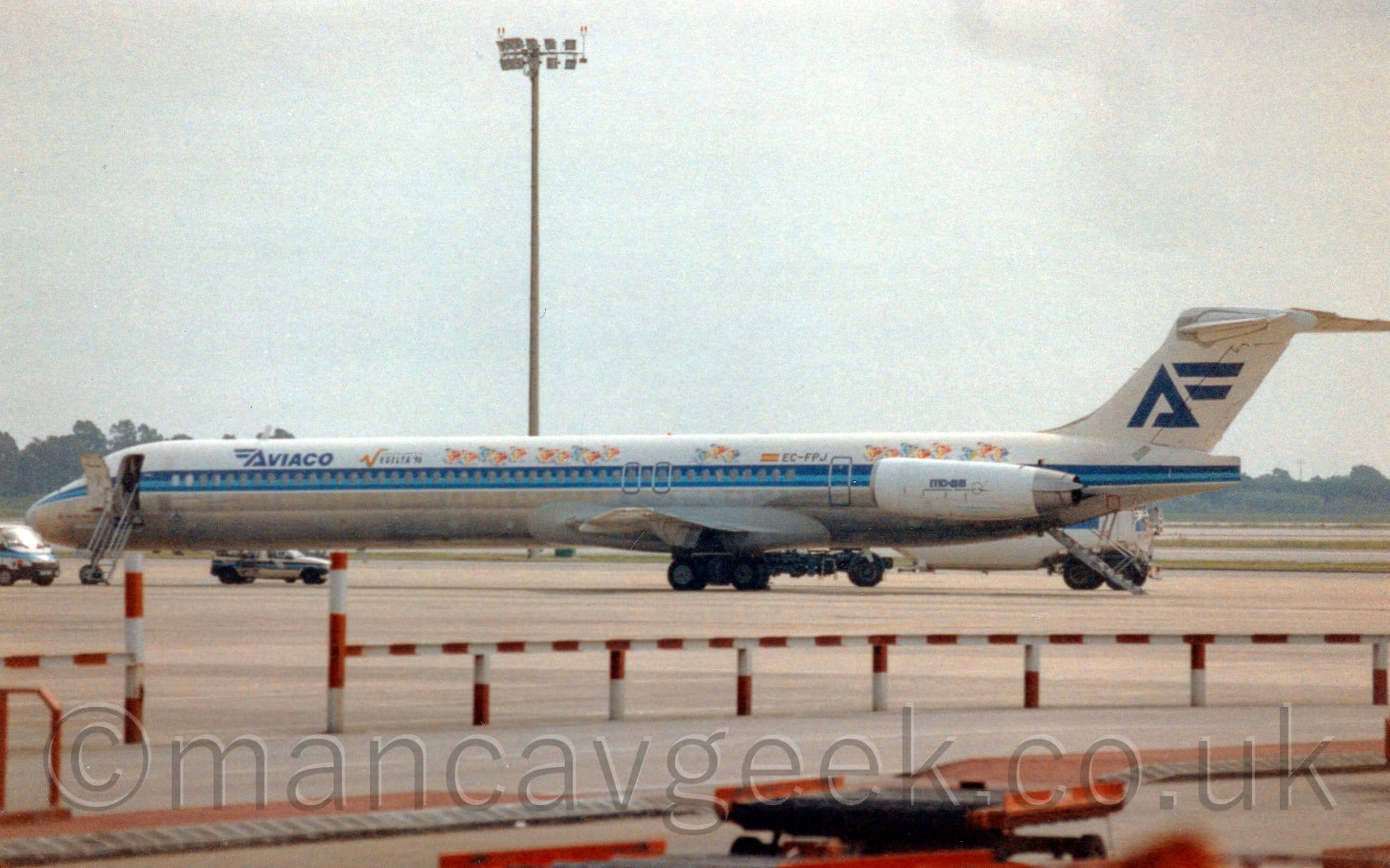 Side view of a twin engined jet airliner with the engines mounted on the sides of the rear fuselage, parked facing to the left, with built-in airstairs extended from the forward door, just aft of the cockpit, and from under the tail, between the engines. The plane is largely white, with a silver belly, and a thick, 2-tone blue stripe running along the body, covering the passenger cabin windows. There are dark blue "Aviaco" titles on the upper forward fuselage, with a pair of stylised wings facing to the left on the initial letter. Almost cartoonish images of bicyclists in various colours adorn the top of the fuselage, along with additional "Vuelta 96" text. There are blue "MD-88" titles on the sides of the white engine pods. The white tail has a larger version of with winged letter "A", this time with the wings facing to the right. Grey concrete apron fills most of the foreground, with a red and white strip[ed metal barrier stretching across most of the frame, with only a small gap towards the left. A tall lighting pole soars into the grim grey sky.