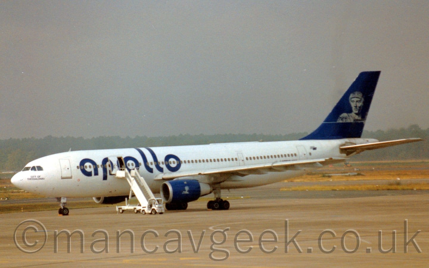 Side view of a twin engined jet airliner parked facing to the left. The plane is mostly white, with large, blue, billboard-style "Apollo" titles on the forward fuselage, blue engine pods under the wings, and a blue tail with a black and white image of a person in vaguely Grecian garb. A white set of mobile air stairs is attached to the mid-cabin door just forward of the wing. Grey concrete apron fills the foreground, with yellowing grass in the background leading off to trees vanishing in to haze in the distance. Flat grey sky fills the frame.